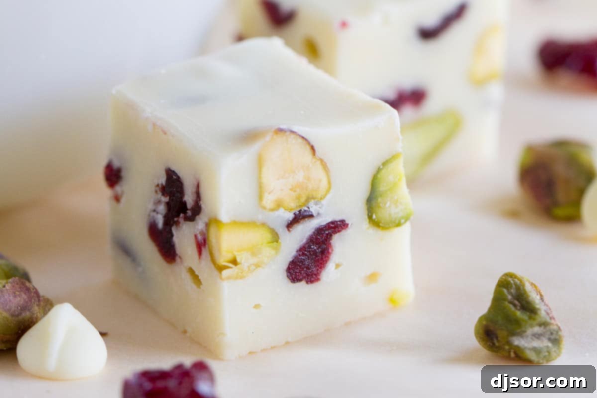 Square of creamy cranberry pistachio fudge surrounded by nuts, cranberries, and a white chocolate chip.