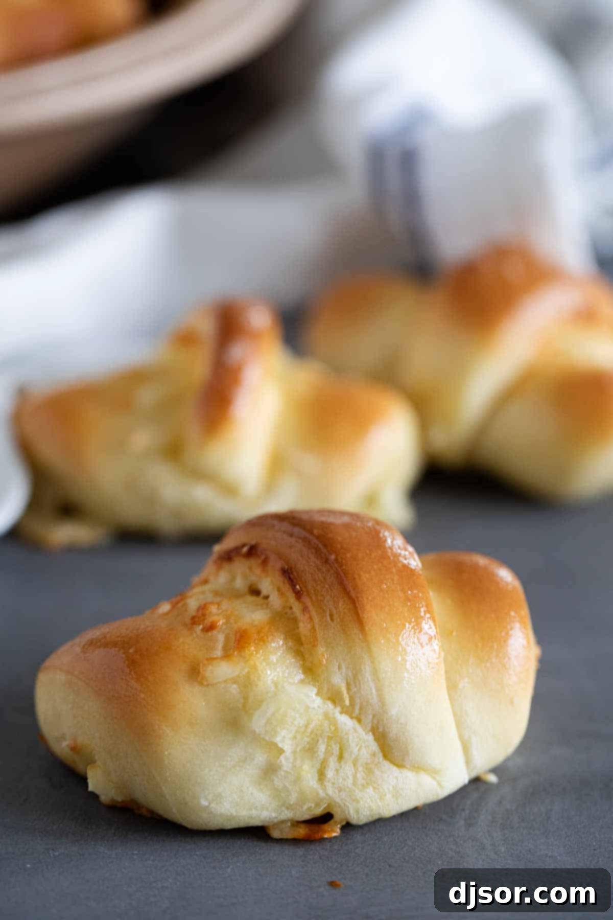 A perfectly golden-brown crescent roll resting on a surface, with more delicious rolls blurred in the background.