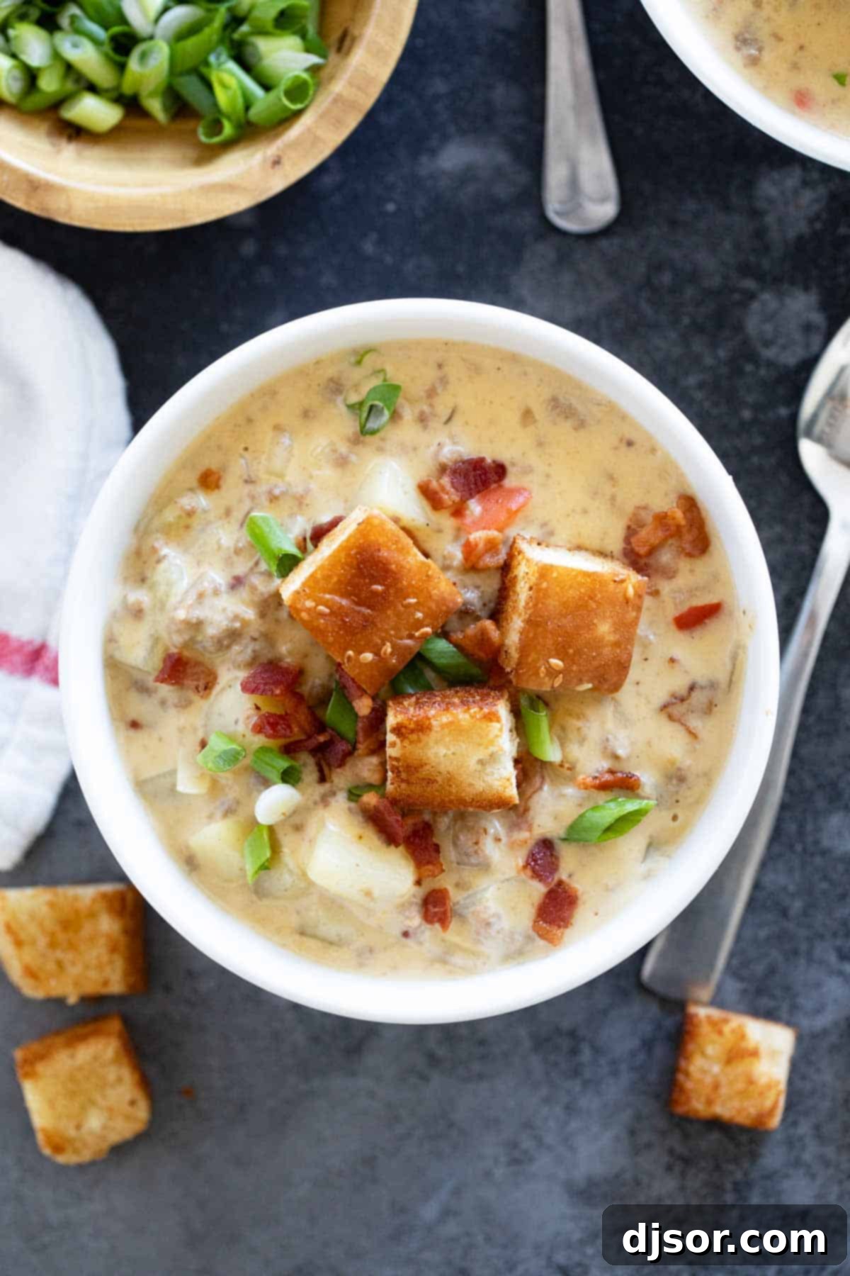 A close-up shot of a steaming bowl of bacon cheeseburger chowder, topped generously with crispy bacon bits, homemade croutons, and fresh green onions, ready to be enjoyed.