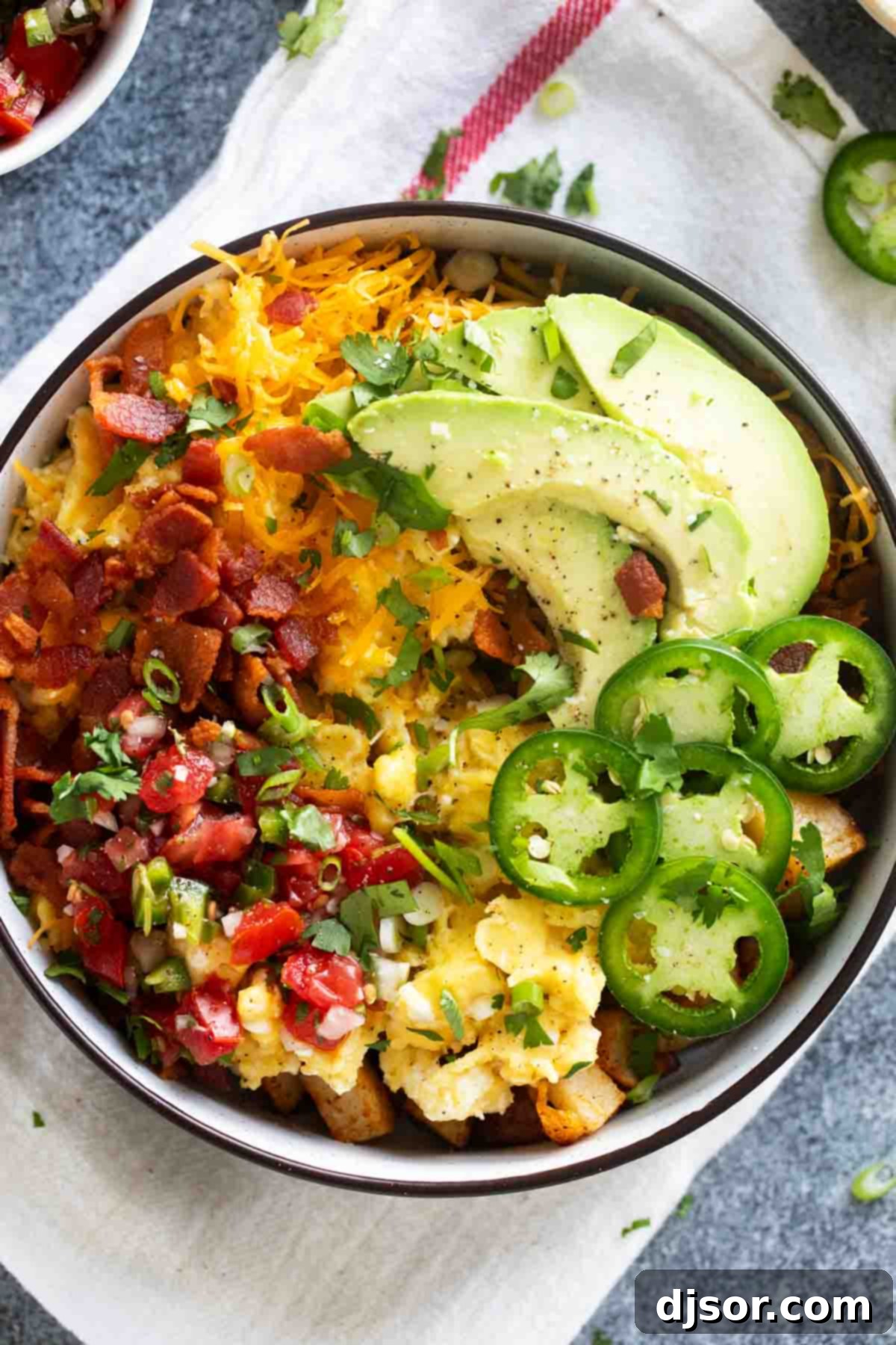 A beautifully assembled breakfast bowl featuring potatoes, scrambled eggs, crispy bacon, sliced avocado, jalapeños, and fresh herbs, ready to eat.