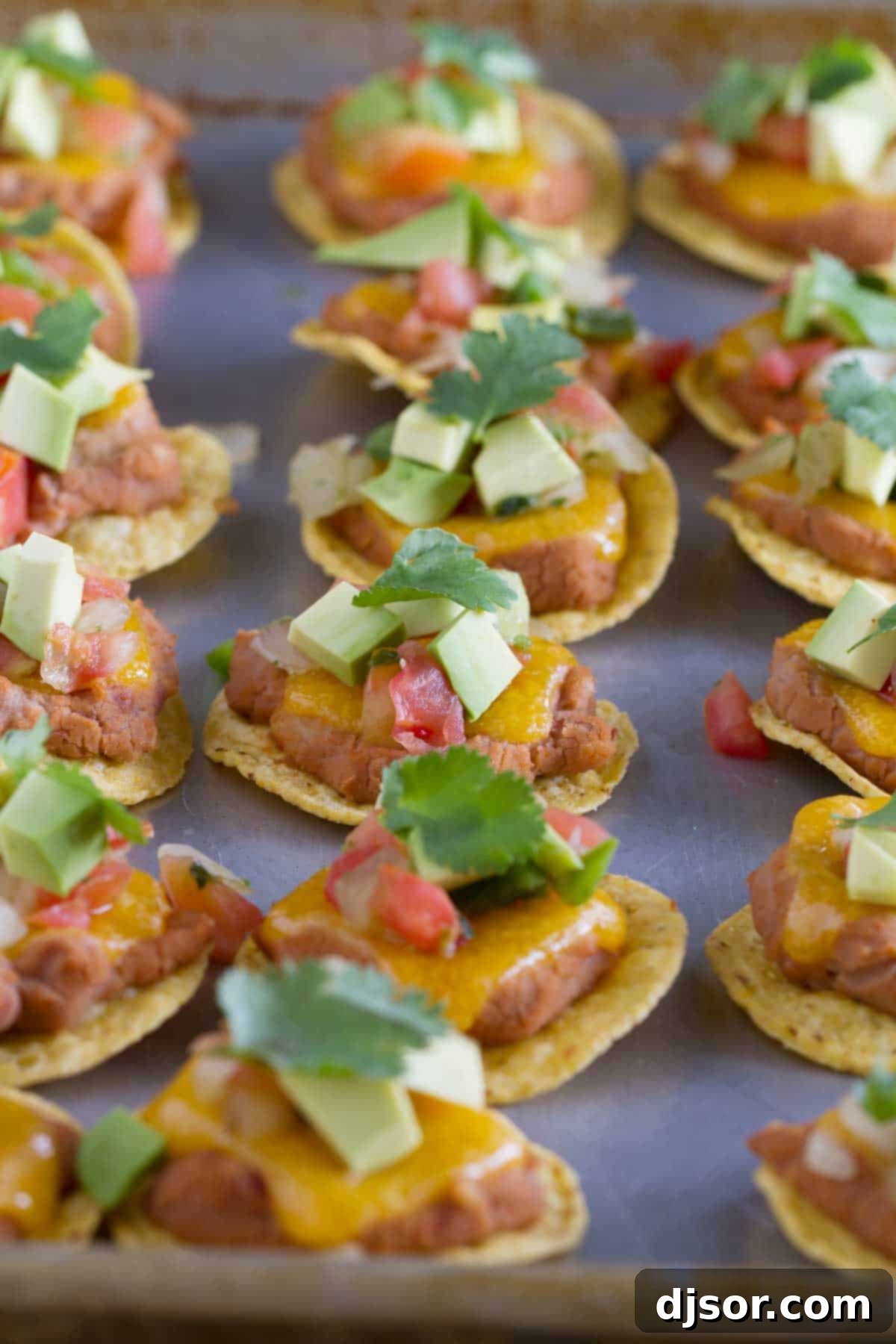 Rows of partially baked Nacho Bites on a baking sheet, with melted cheese over refried beans on tortilla chips.