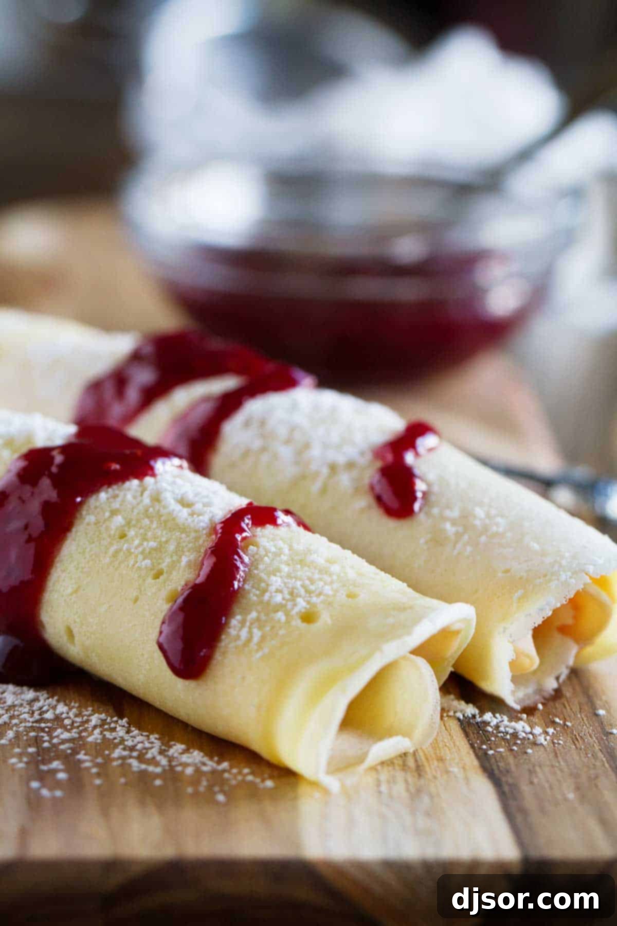 Two perfectly rolled Monte Cristo Crepes, golden and inviting, garnished with a vibrant raspberry jam and a light dusting of powdered sugar, resting on a rustic cutting board.