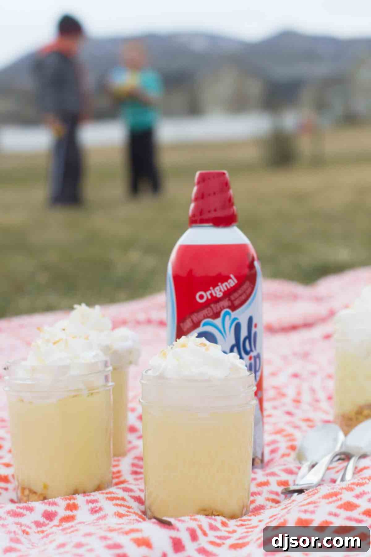 Three jars of Coconut Lemon Pudding Parfait sit invitingly on a picnic blanket, accompanied by a can of Reddi-wip, ready for a delightful outdoor treat.