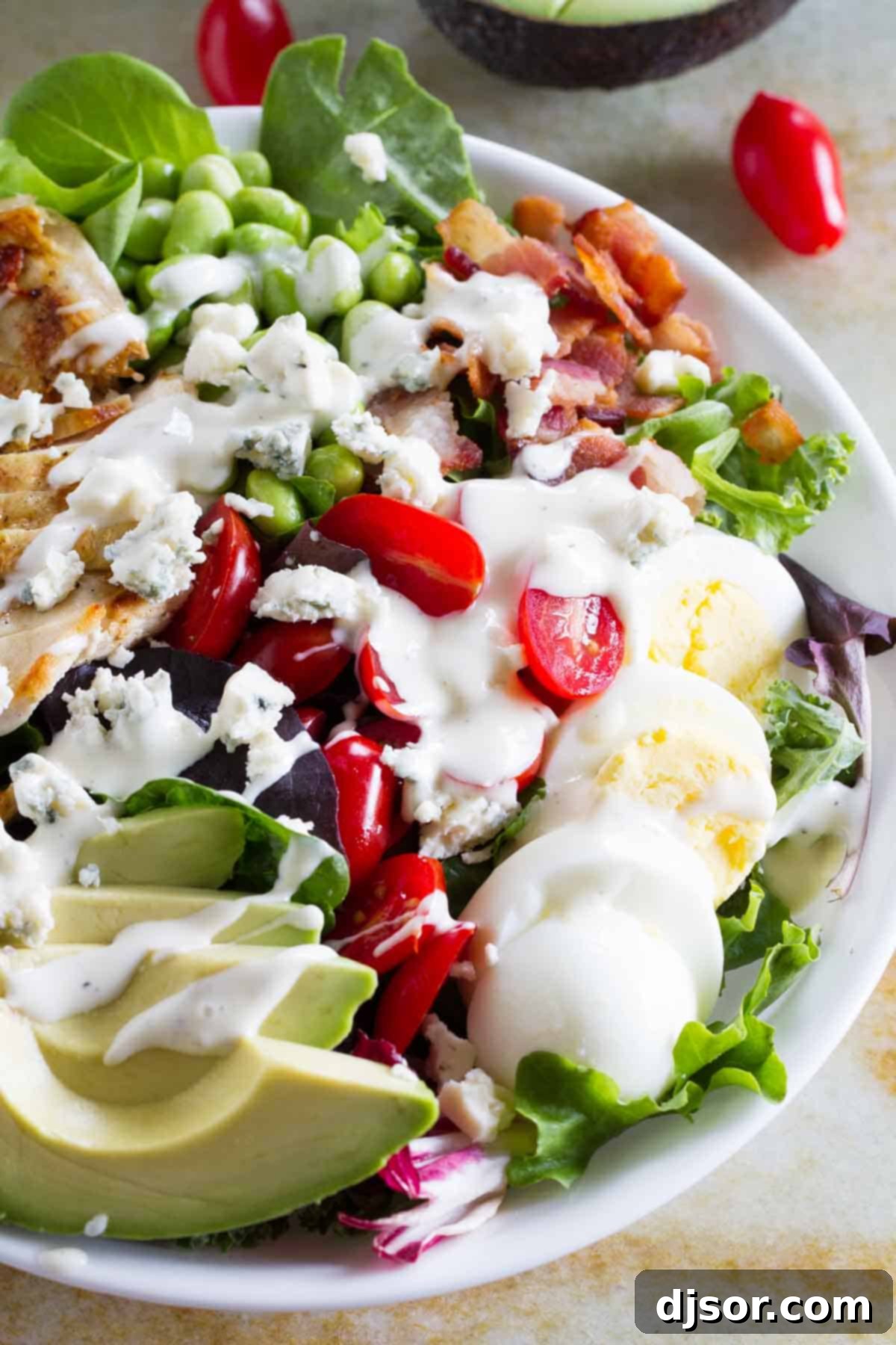 A top-down view of a beautifully assembled Protein-Packed Cobb Salad, showcasing slices of grilled chicken, crispy bacon, avocado, tomatoes, edamame, hard-boiled eggs, and crumbled blue cheese, drizzled with creamy ranch dressing.