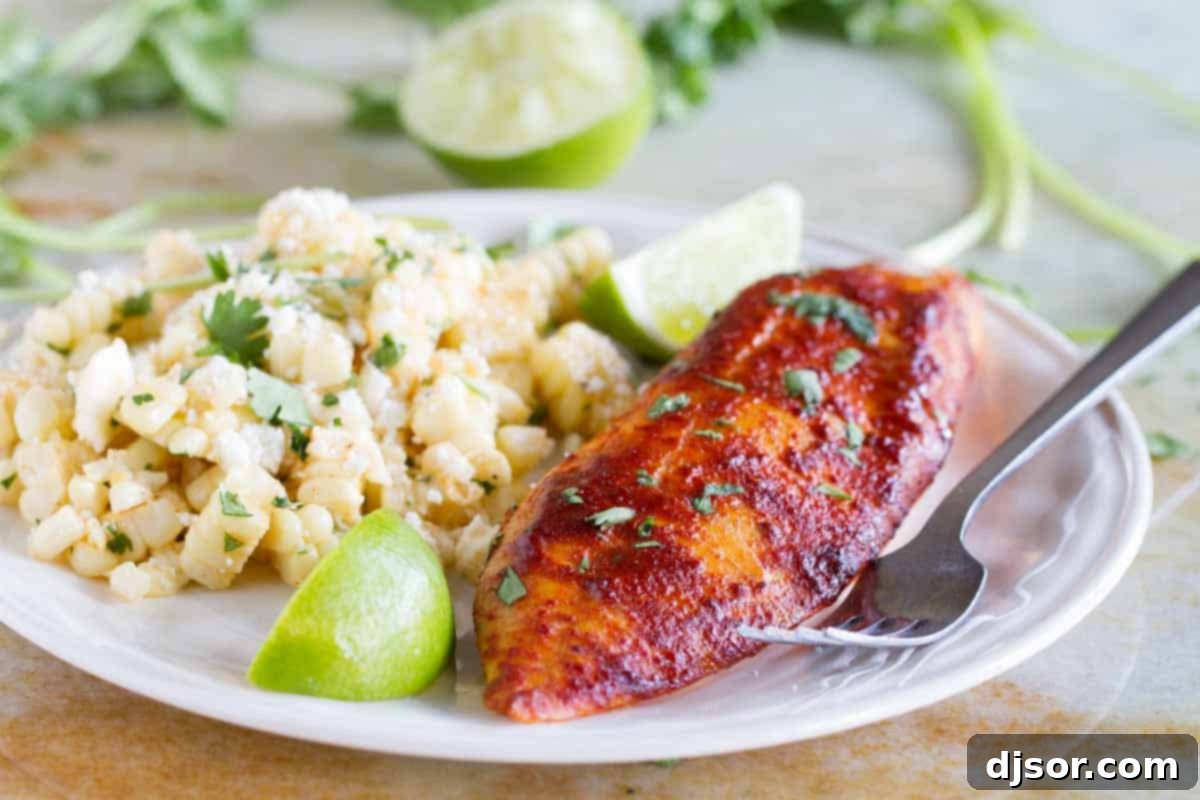 A white plate featuring a serving of taco seasoned grilled fish, accompanied by a vibrant Mexican corn salad and fresh lime wedges, highlighting a complete meal.
