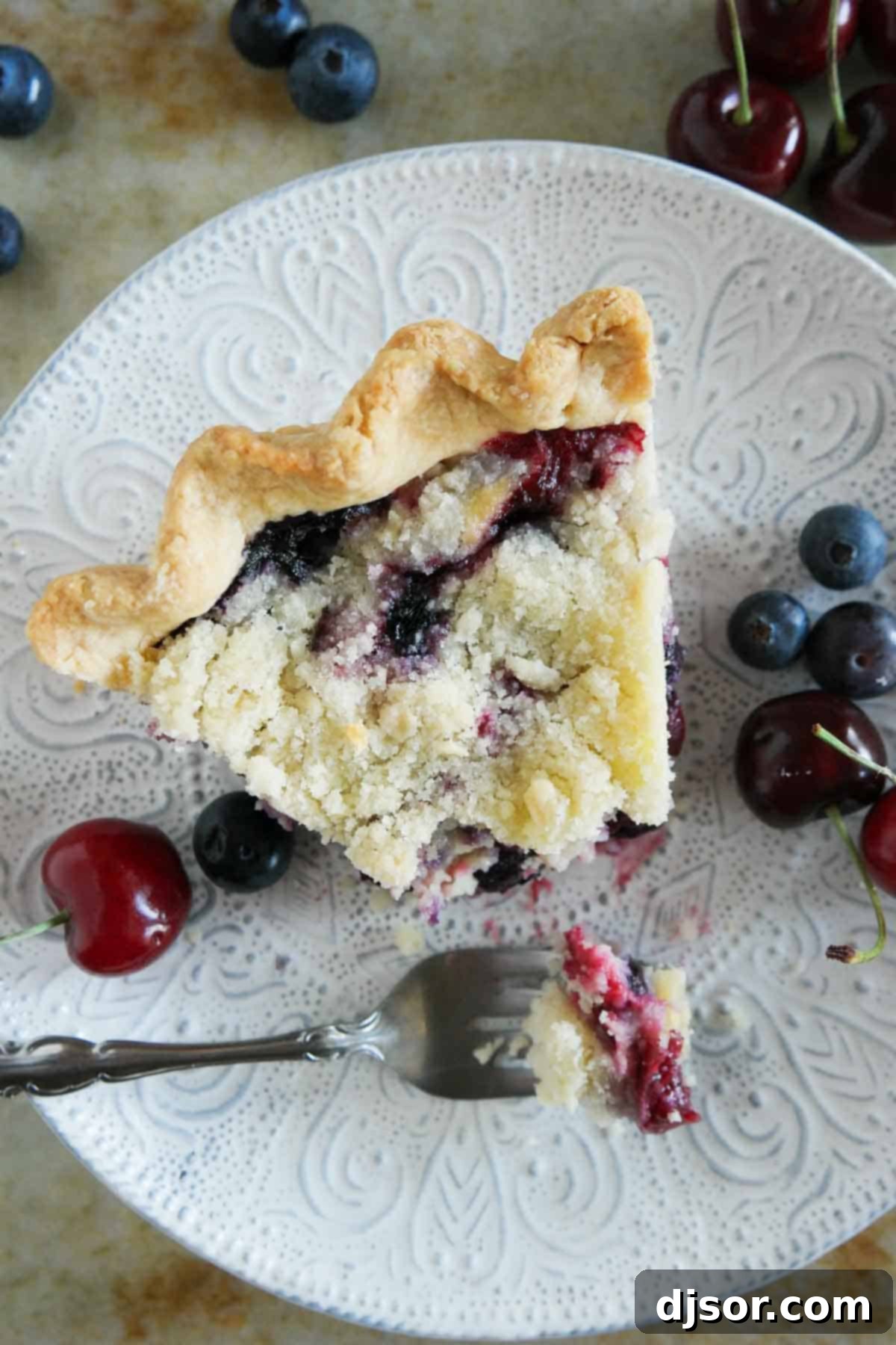 A close-up of a slice of Cherry and Blueberry Cream Pie on a white plate, with a fork having taken a bite, revealing the rich layers of fruit, cream, and streusel.