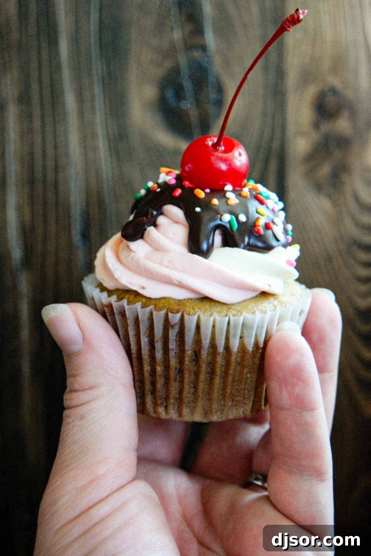A hand presenting a single banana split cupcake, showcasing its delicious layers of frosting and toppings