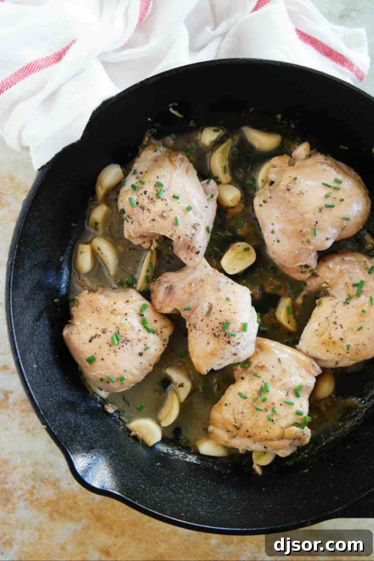 Close-up of a cast iron skillet filled with golden-brown chicken thighs, fragrant garlic cloves, and fresh herbs.