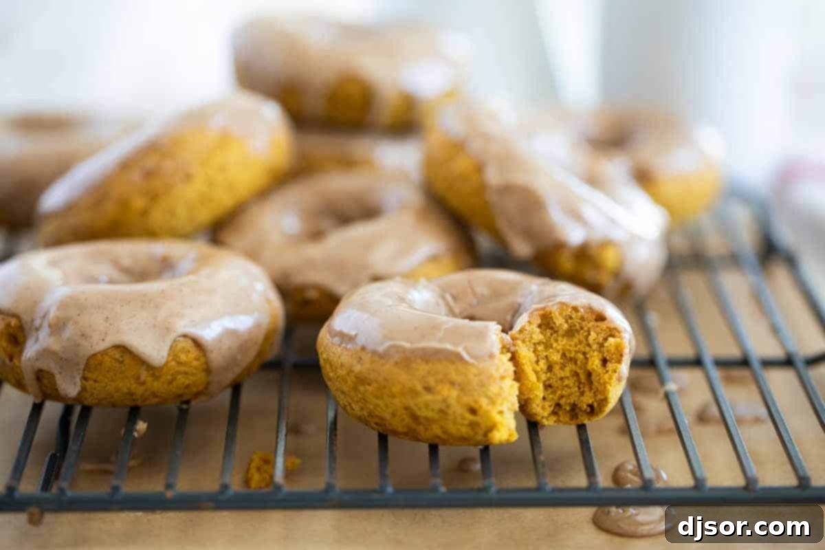 A close-up of a baked pumpkin donut on a cooling rack with a bite taken out, revealing its moist, spiced interior and thick maple cinnamon glaze.