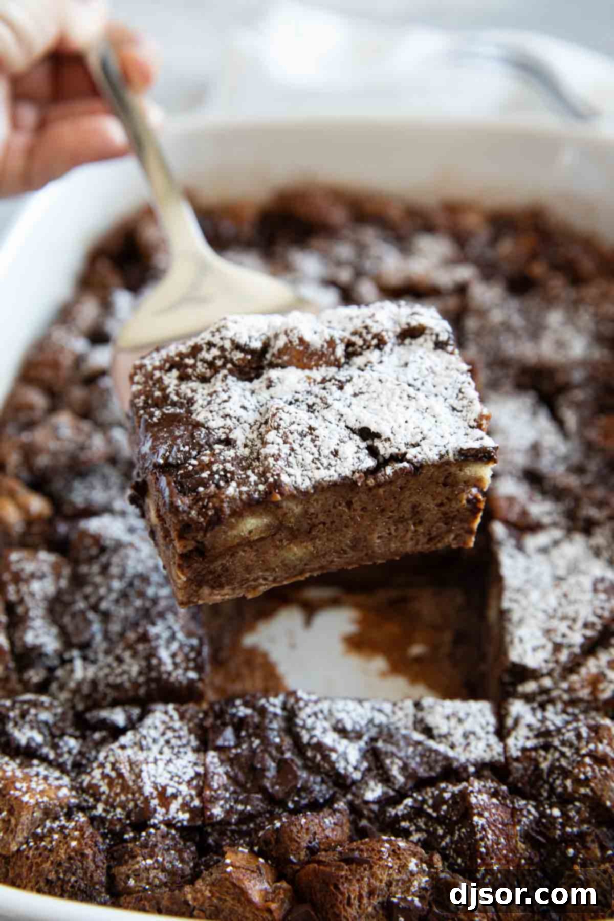 A close-up shot of a spoon scooping a generous portion of warm, gooey chocolate bread pudding from a baking dish.