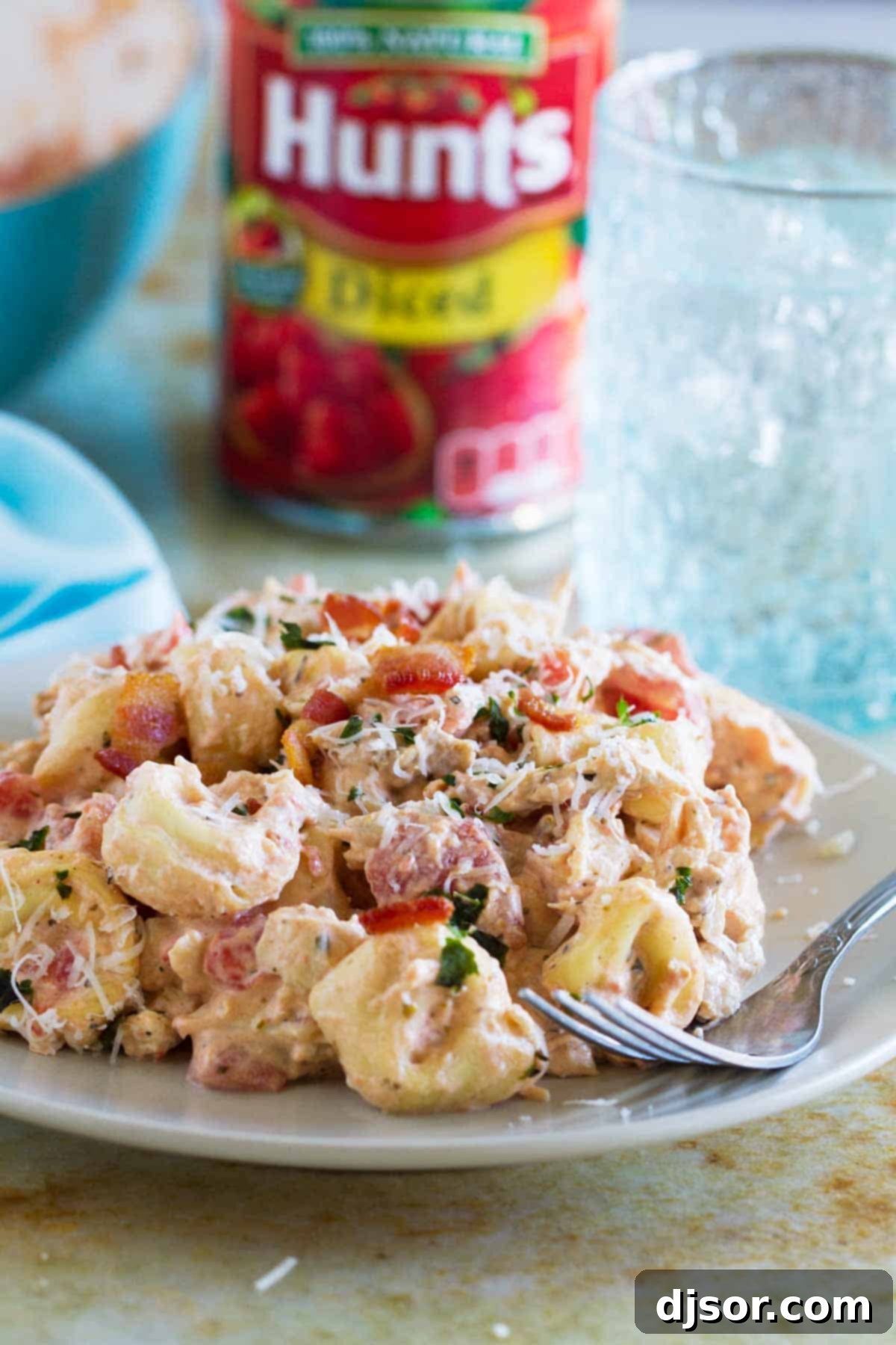 A close-up of a plate filled with Creamy Tomato Tortellini with Chicken and Bacon, with a can of Hunt's Diced Tomatoes subtly blurred in the background, beautifully highlighting a key ingredient and the freshness it brings to the dish.