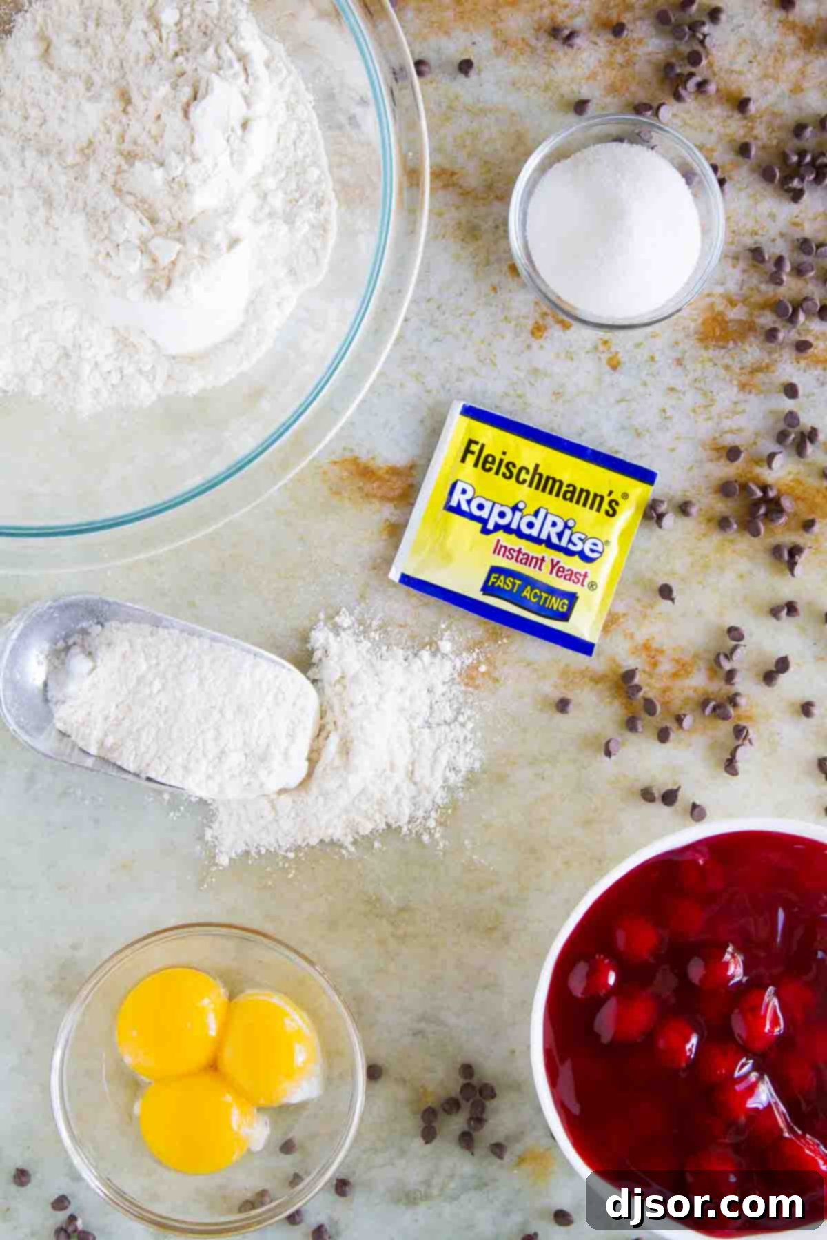 A selection of fresh ingredients laid out, including flour, sugar, yeast, milk, butter, eggs, cherry pie filling, and mini chocolate chips, ready for baking Chocolate Cherry Rolls.