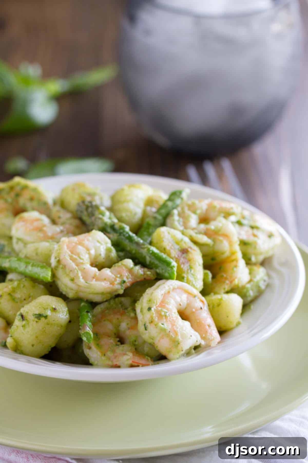 Vibrant Pesto Gnocchi with Shrimp and Asparagus 2 A beautifully plated dish of gnocchi tossed in vibrant green pesto, complemented by succulent pink shrimp and crisp green asparagus pieces. Garnished with fresh basil leaves and a sprinkle of grated Parmesan cheese, ready to be served.