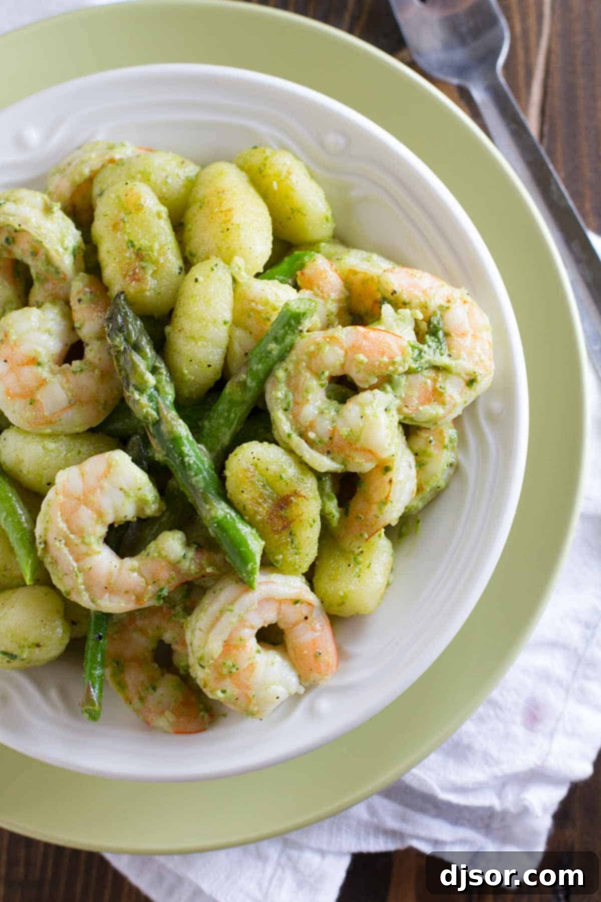 Vibrant Pesto Gnocchi with Shrimp and Asparagus 3 A generous serving of Gnocchi with Pesto, Shrimp, and Asparagus, presented in a shallow, wide bowl. The vibrant green pesto coats each gnocchi, with plump pink shrimp and bright green asparagus spears interspersed throughout. A fork is resting in the bowl, ready to enjoy.