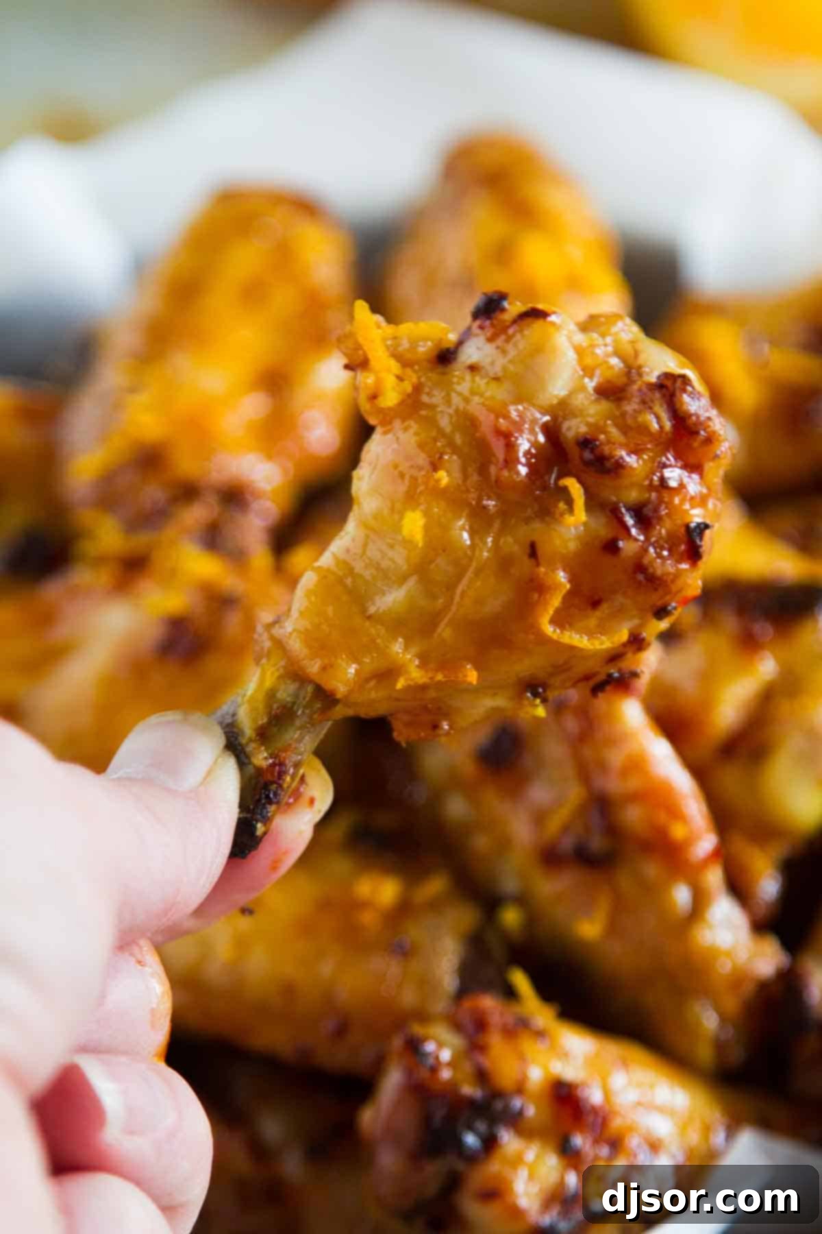 A hand holding a single glistening Chipotle Orange Chicken Wing, with a blurred background of more wings in a bowl.