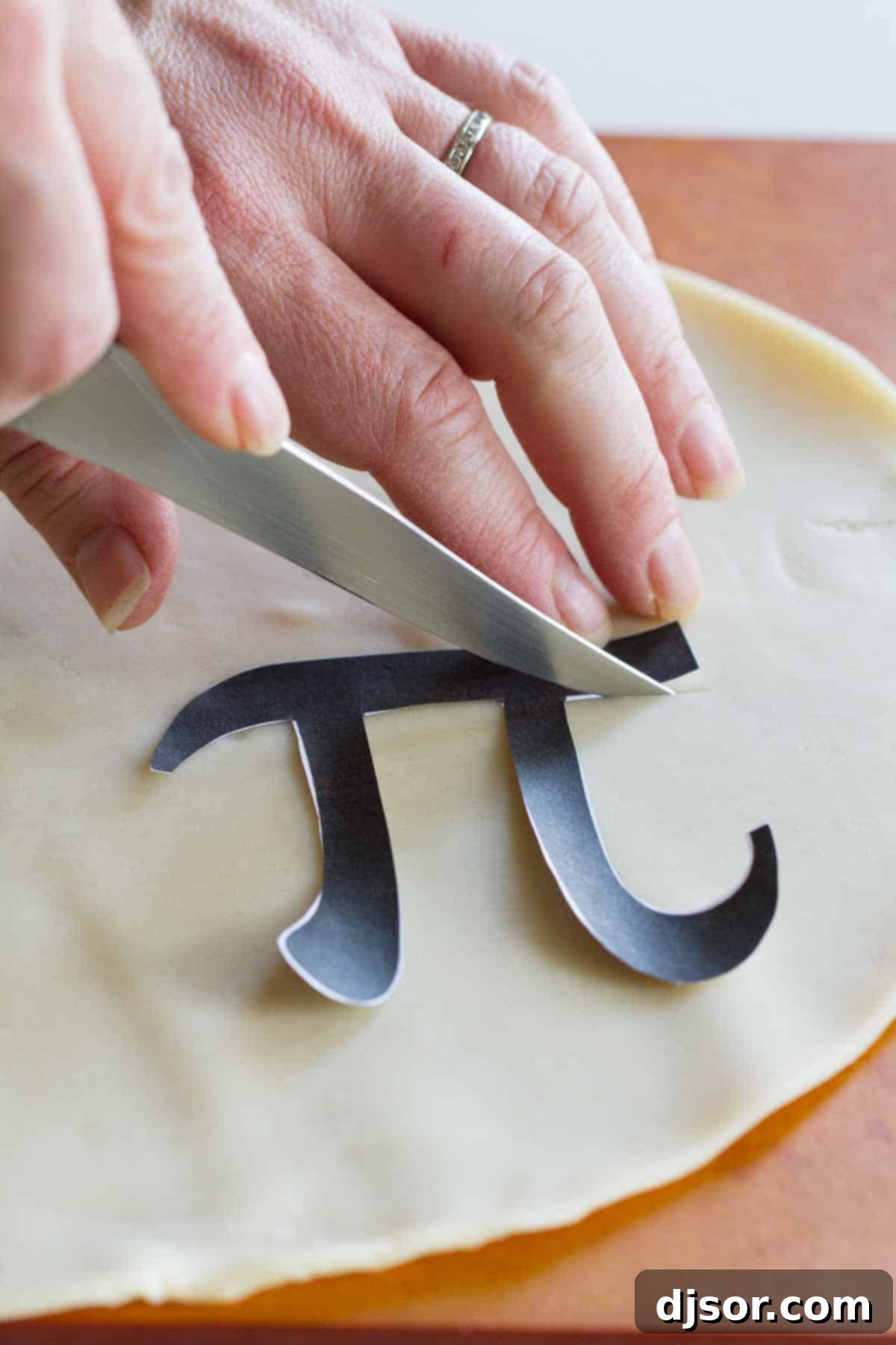 Hands carefully cutting out a 'Pi' symbol from extra pie dough using a stencil, for decorative placement on a pie.