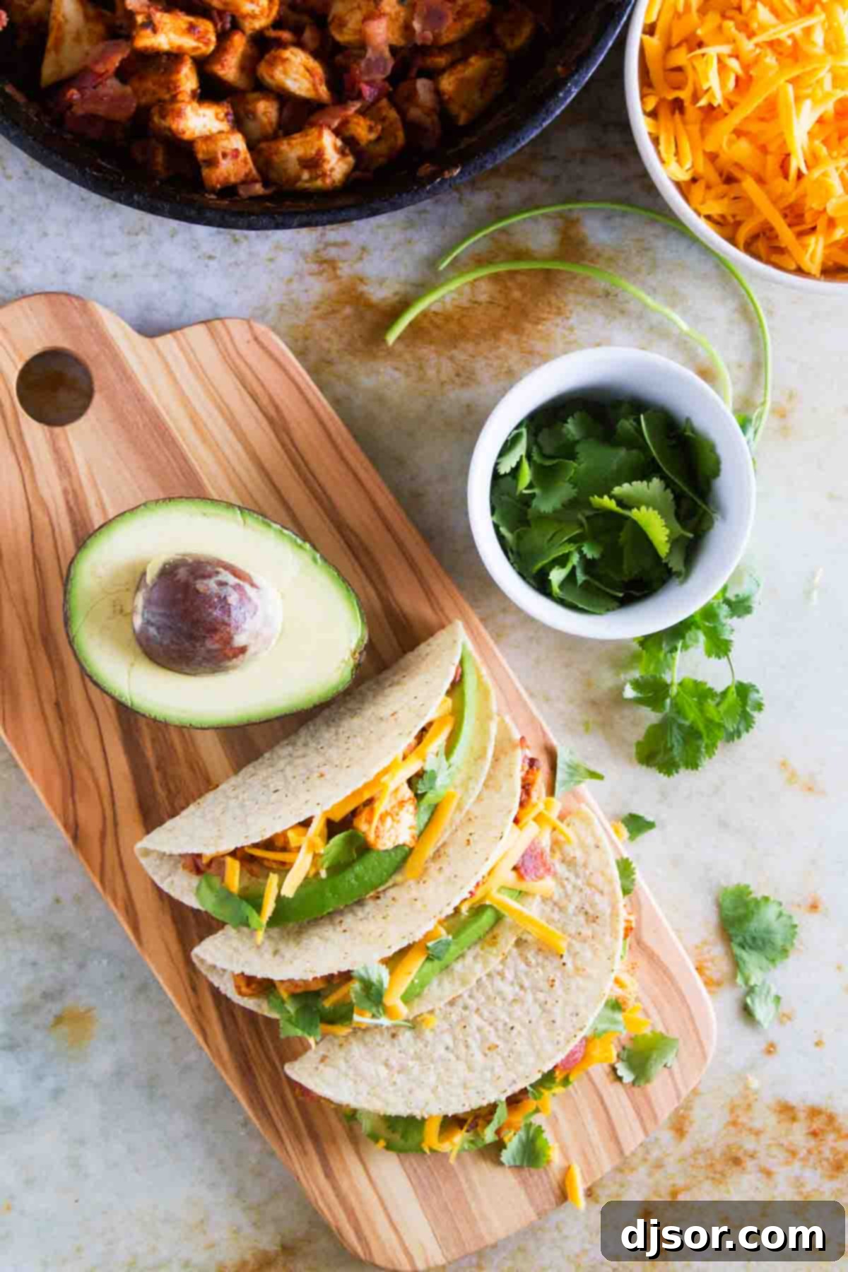 Stacked Chicken Bacon Avocado Tacos 6 Several Chicken Bacon Avocado Tacos arranged on a cutting board, garnished with fresh cilantro, avocado slices, and shredded cheddar cheese for a vibrant presentation.