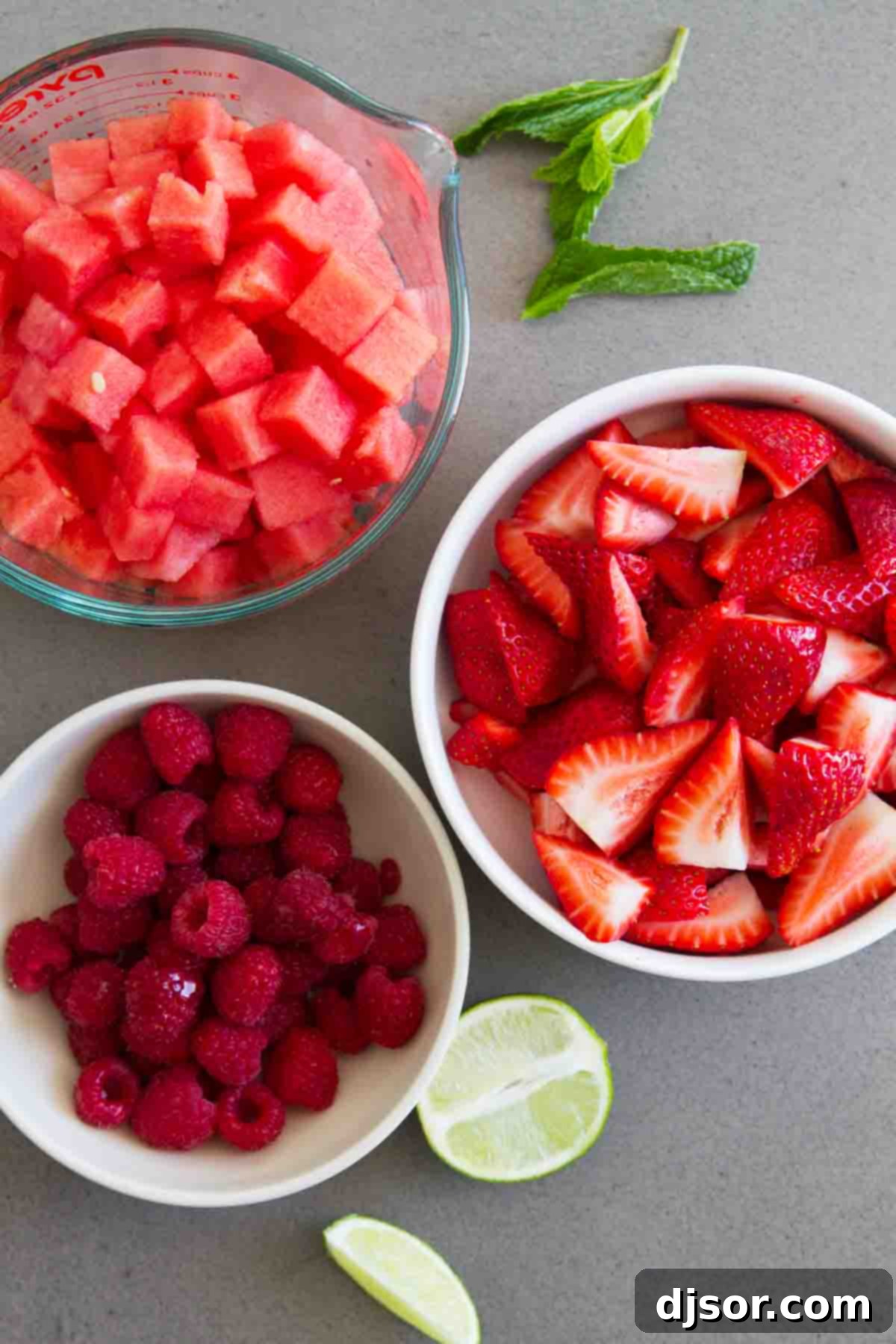 Gathering Ingredients for Crown Ruby Fruit Salad - Simple and Fresh Individual ingredients for Crown Ruby Fruit Salad laid out: granulated sugar, water, lime juice, lime zest, cubed watermelon, quartered strawberries, a package of raspberries, and chopped mint leaves.