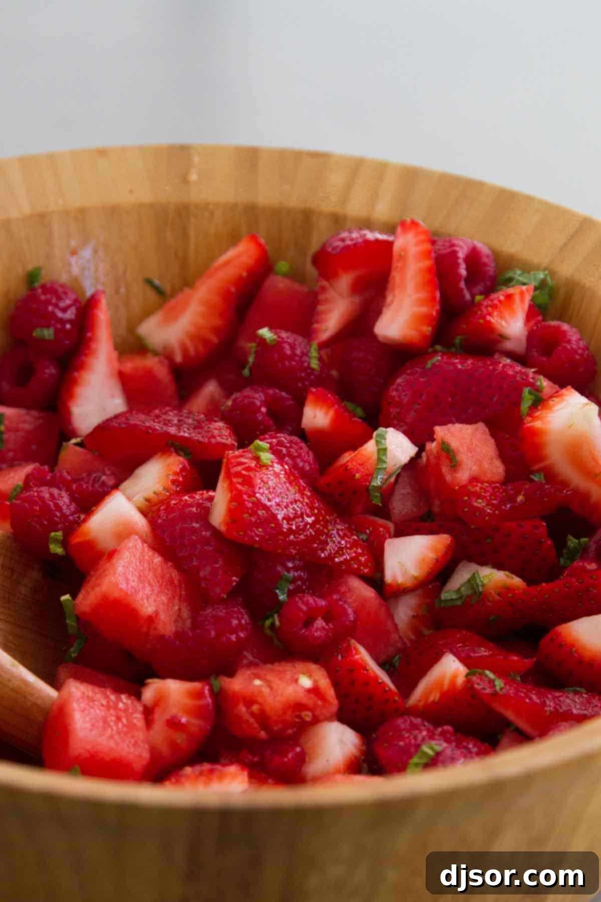 Crown Ruby Fruit Salad in Wooden Bowl - A Refreshing Summer Treat A rustic wooden bowl brimming with Crown Ruby Fruit Salad, showcasing a vibrant mix of red watermelon, strawberries, and raspberries, lightly glistening with lime syrup and dotted with fresh mint.