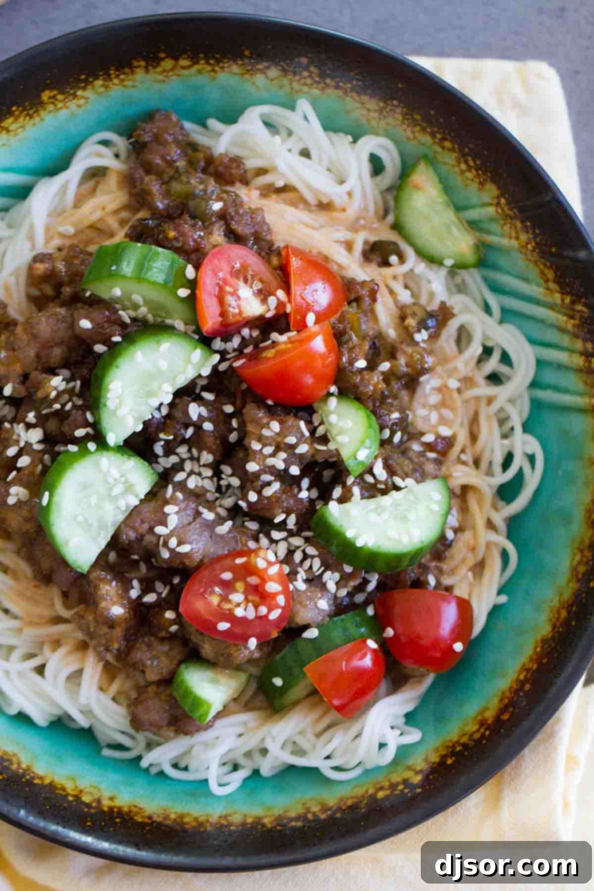 Top view of Spicy Pork Noodle Bowl garnished with sliced cucumbers, grape tomatoes, and sesame seeds.