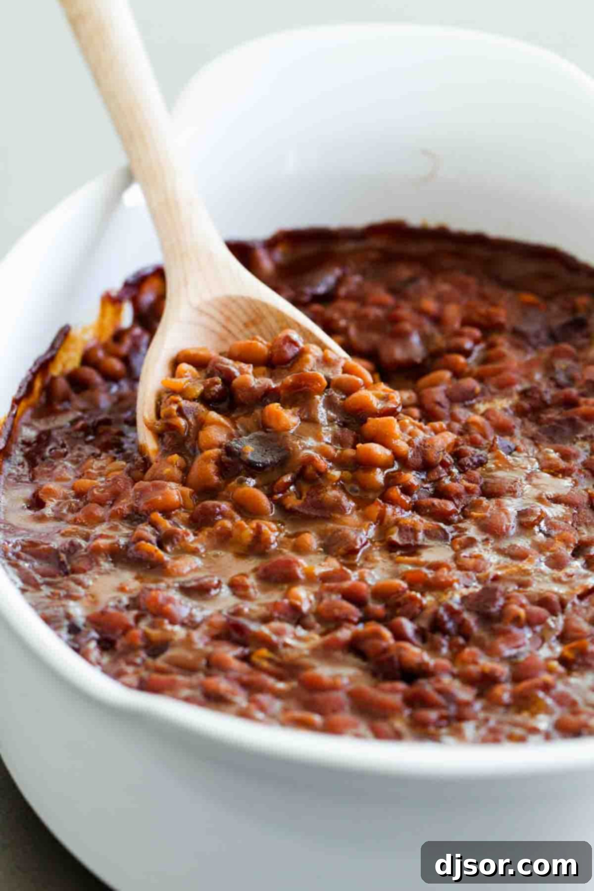 Baked beans with bacon in a casserole dish with a wooden spoon to scoop, highlighting the rich texture and crispy bacon bits.