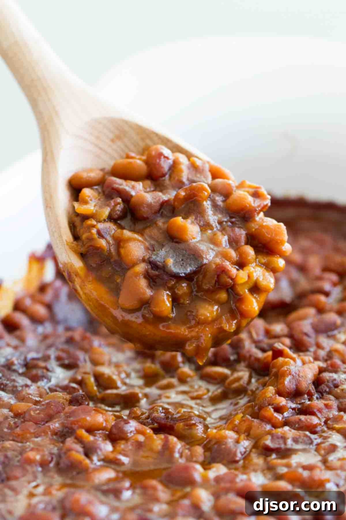 A wooden spoon scooping a portion of rich, saucy baked beans with visible pieces of bacon, ready to be served.