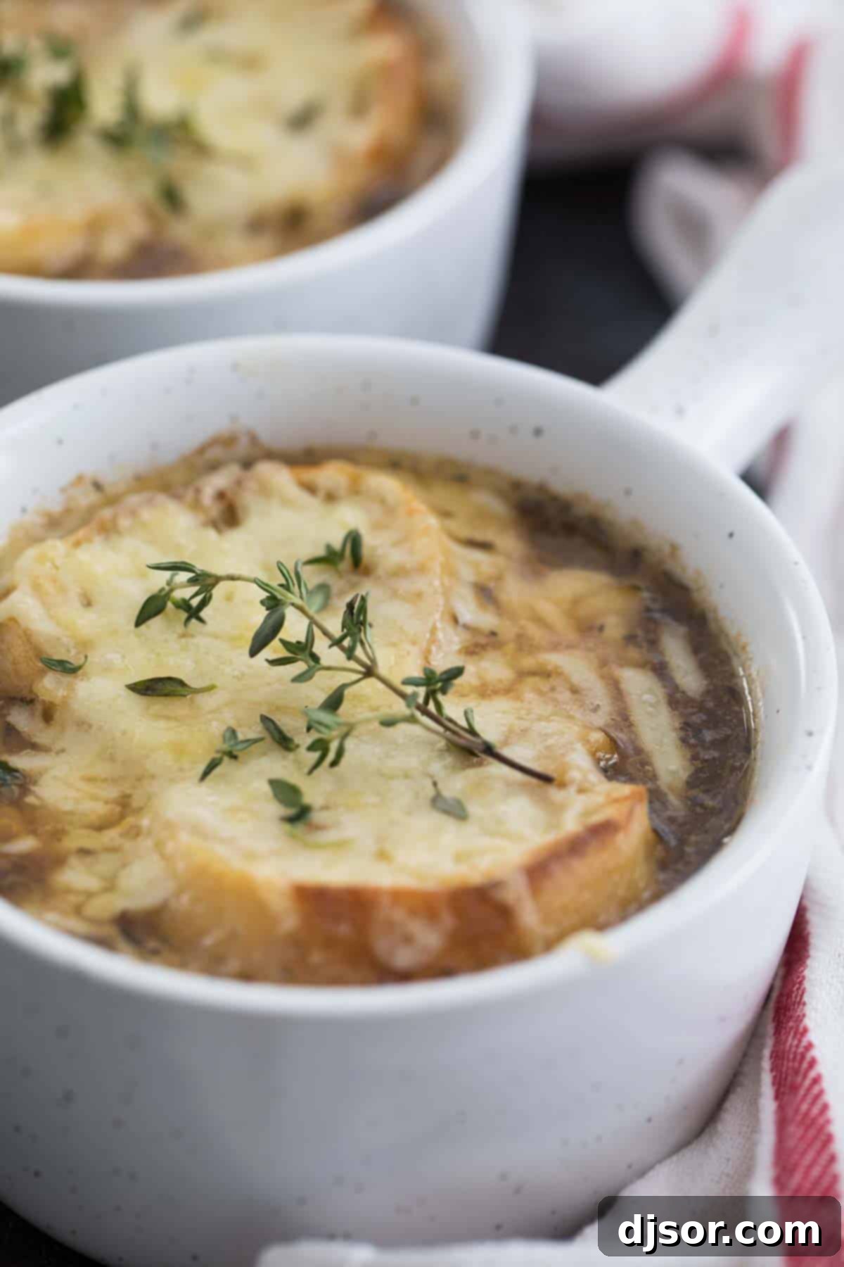 A close-up shot of French onion soup in an oven-safe bowl, with a thick layer of melted, browned Gruyere cheese and a sprig of fresh thyme