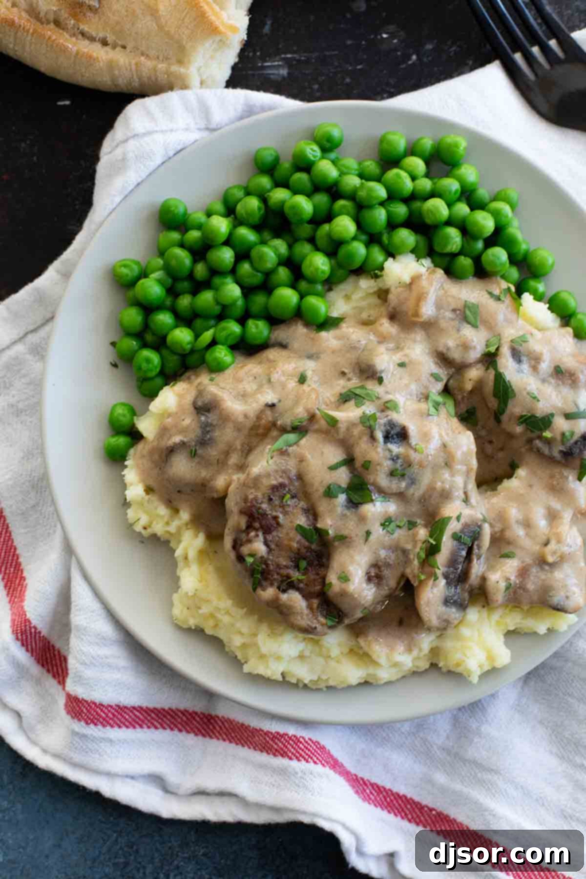 An inexpensive, filling and comforting meal that is family friendly and full of flavor, this Salisbury Steak Recipe is a childhood throwback that you’ll want to make over and over again. A plate featuring Salisbury steak with gravy over mashed potatoes and a side of green peas.