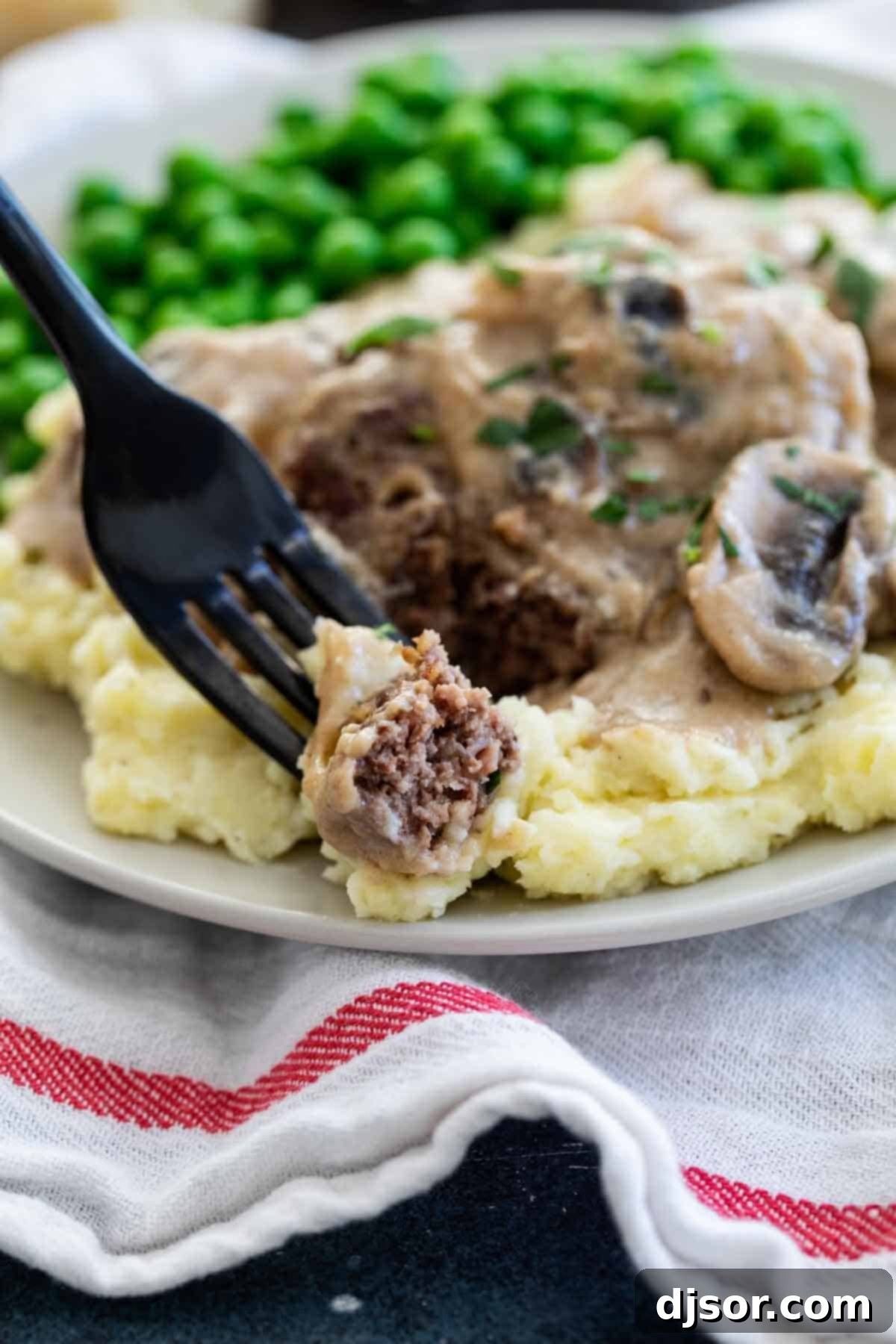An inexpensive, filling and comforting meal that is family friendly and full of flavor, this Salisbury Steak Recipe is a childhood throwback that you’ll want to make over and over again. A fork holding a bite of Salisbury steak with gravy, showing the delicious texture.