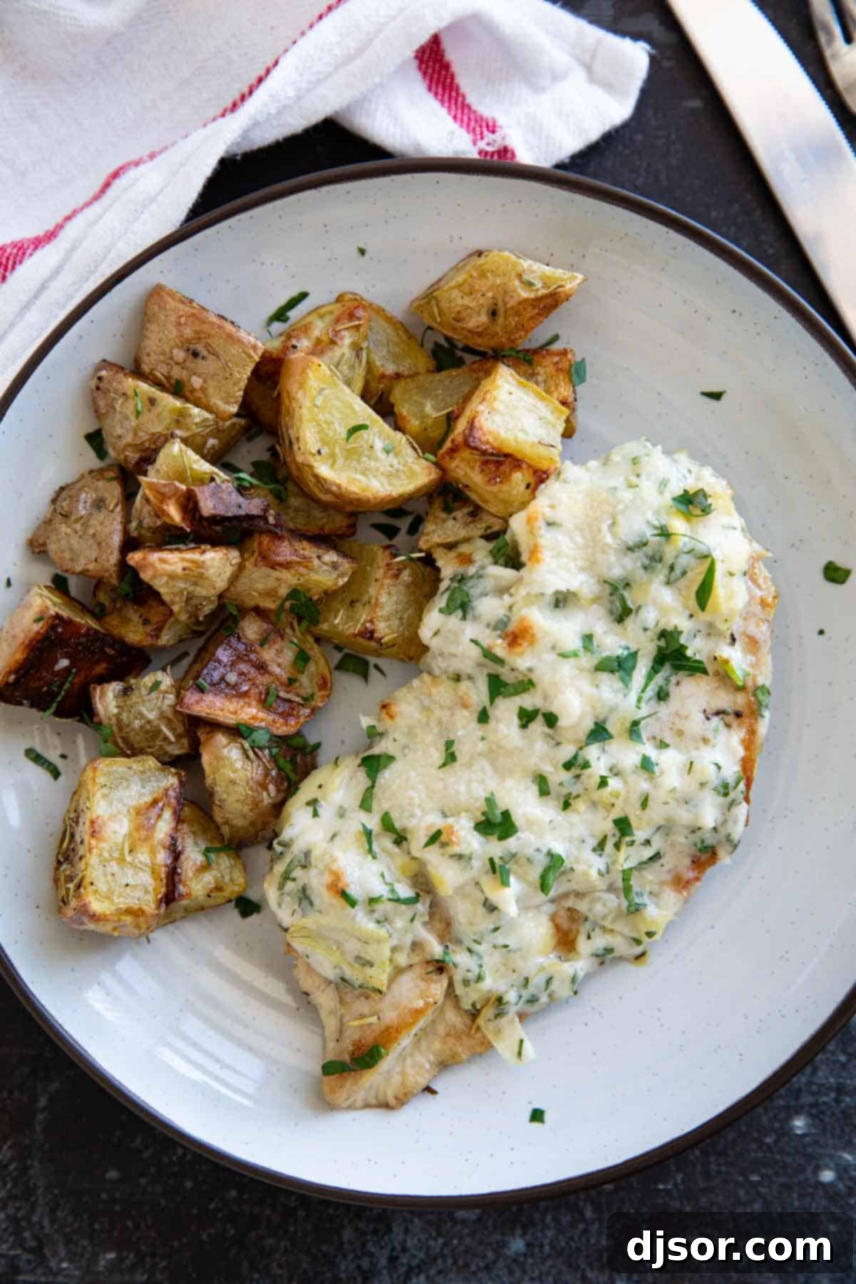 Creamy Lemon Artichoke Chicken 7 A single serving of Artichoke Chicken on a white plate, topped with a rich, melted artichoke and Parmesan crust, accompanied by crispy roasted potatoes and garnished with fresh parsley.