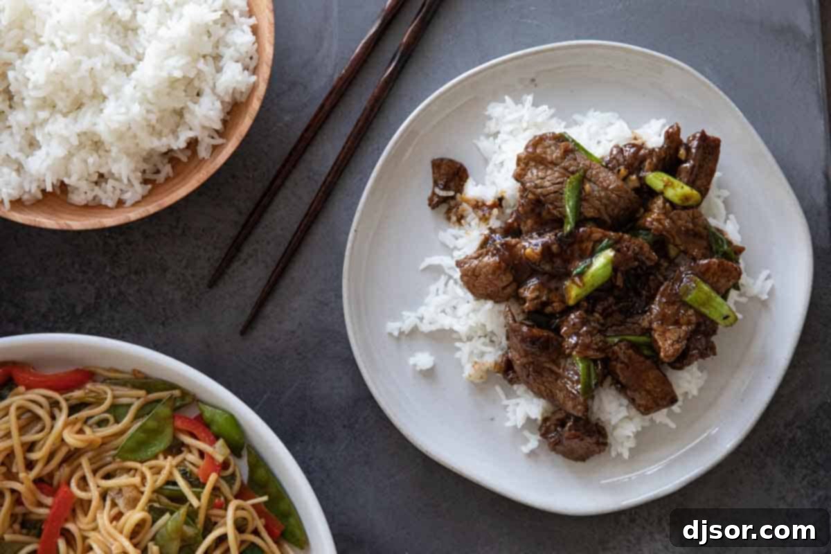 Overhead view of a serving of Mongolian beef over rice on a white plate, with a side of lo mein noodles and a glass of water.