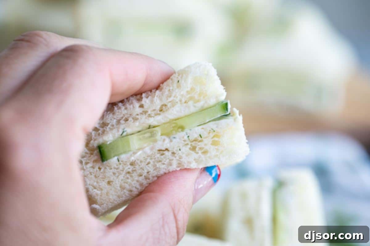 A person holding a single cucumber finger sandwich cut into a triangle, showcasing the creamy dill spread and fresh cucumber slices.