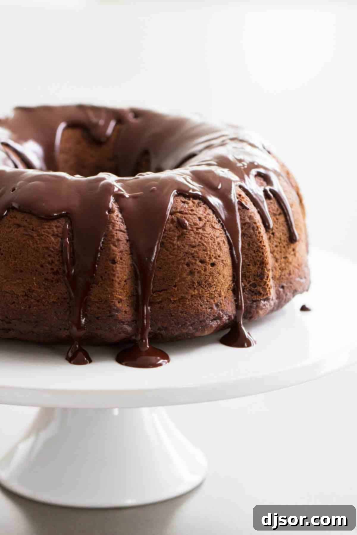 Easy Chocolate Bundt Cake with Satiny Glaze Full chocolate bundt cake with shiny chocolate icing on a white cake stand, ready to be served.
