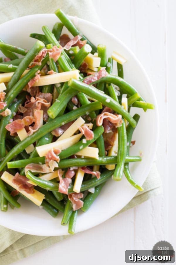 Close-up shot of vibrant green beans coated in a tangy dressing, topped with savory frizzled prosciutto and delectable Gouda cheese pieces.