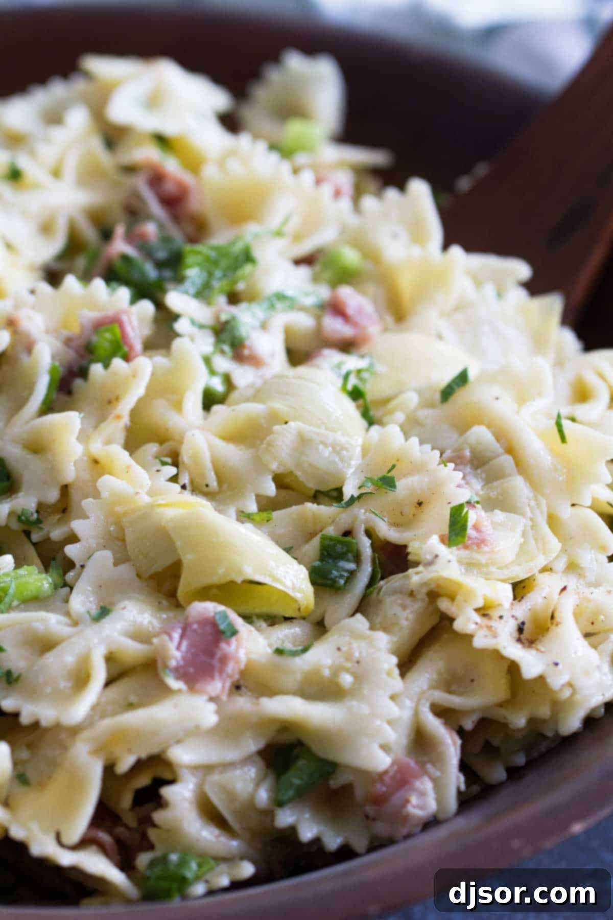 Close-up of Artichoke Pasta Salad, showcasing bowtie pasta, chopped artichokes, bits of prosciutto, and fresh green herbs lightly coated in dressing.