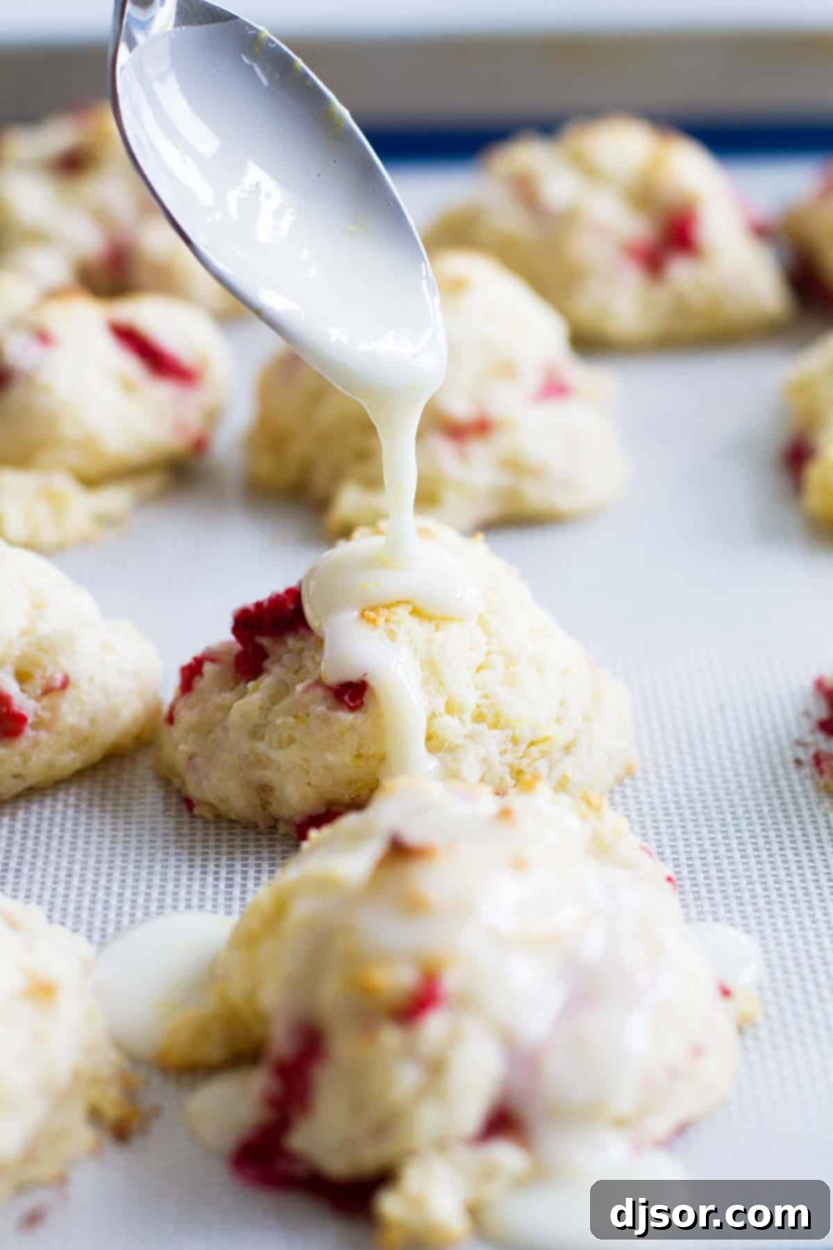 Adding the perfect lemon glaze to warm Raspberry Lemon Breakfast Biscuits. Close-up of a hand pouring the tangy lemon glaze over freshly baked Raspberry Lemon Breakfast Biscuits on a cooling rack.