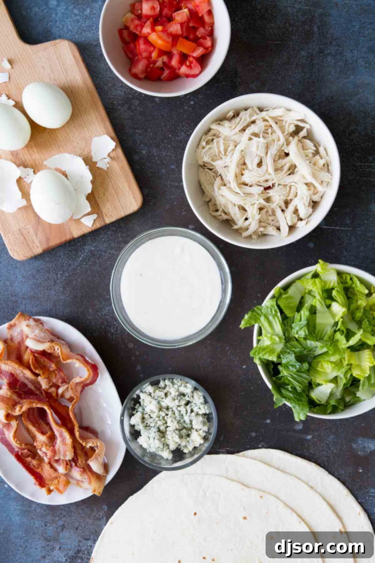 A selection of fresh ingredients laid out for Chicken Cobb Salad Wraps, including shredded chicken, bacon, eggs, lettuce, and tomatoes.
