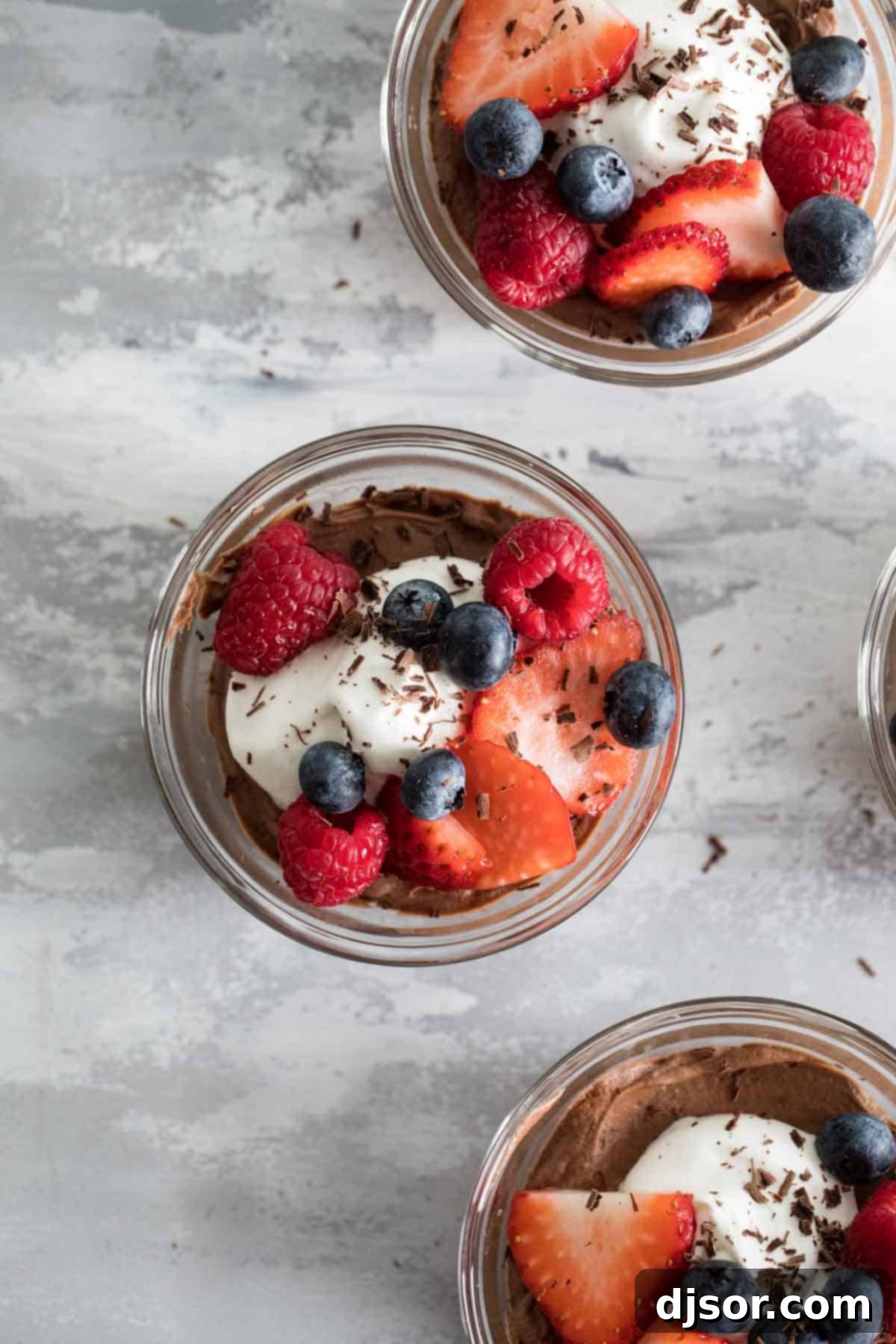 Three individual serving bowls filled with dark chocolate mousse, topped generously with fresh whipped cream and a mix of colorful berries like raspberries, blueberries, and blackberries.