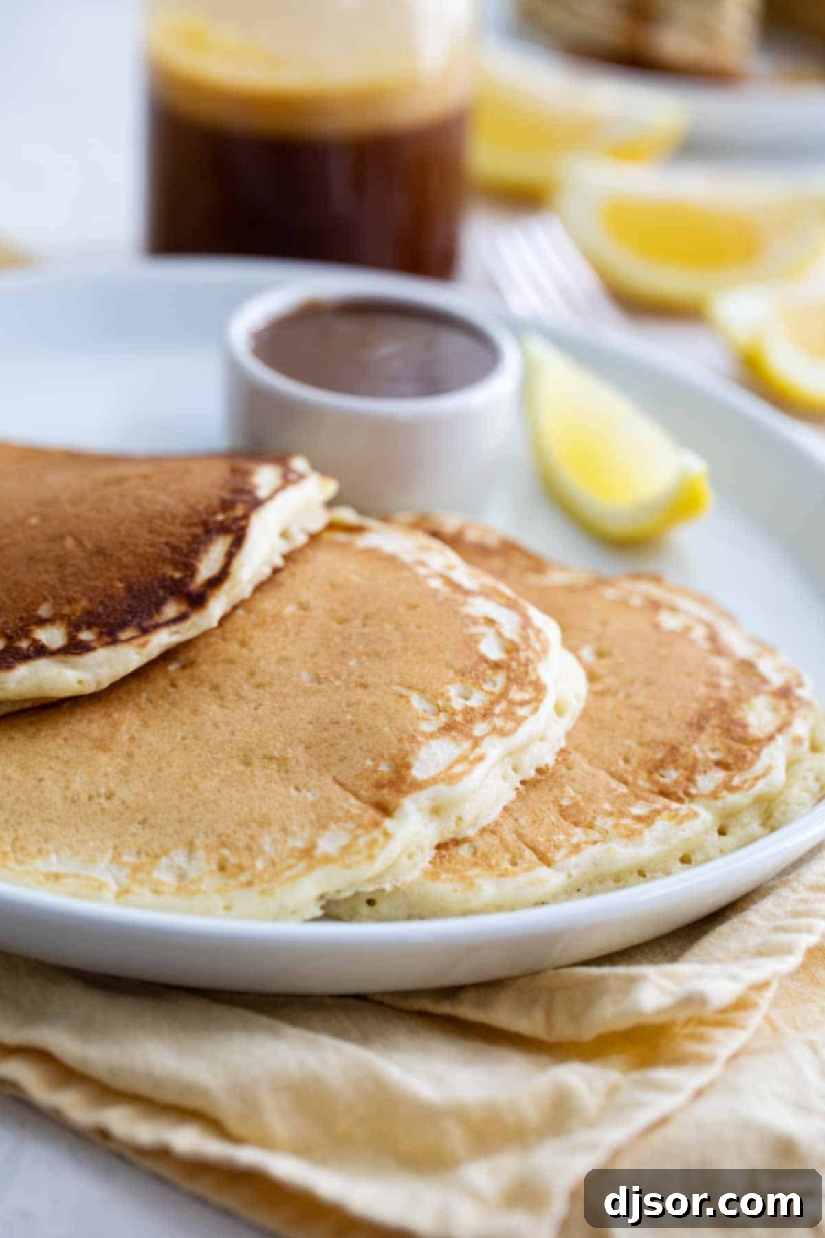 A stack of golden Lemon Pancakes on a white plate, garnished with a fresh lemon slice and a dusting of powdered sugar, ready to be served.