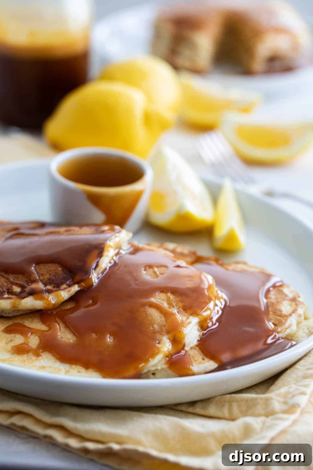 A beautifully composed shot of fluffy Lemon Pancakes, generously drizzled with golden Buttermilk Syrup, alongside fresh lemon halves and whole lemons, emphasizing the vibrant citrus flavor.