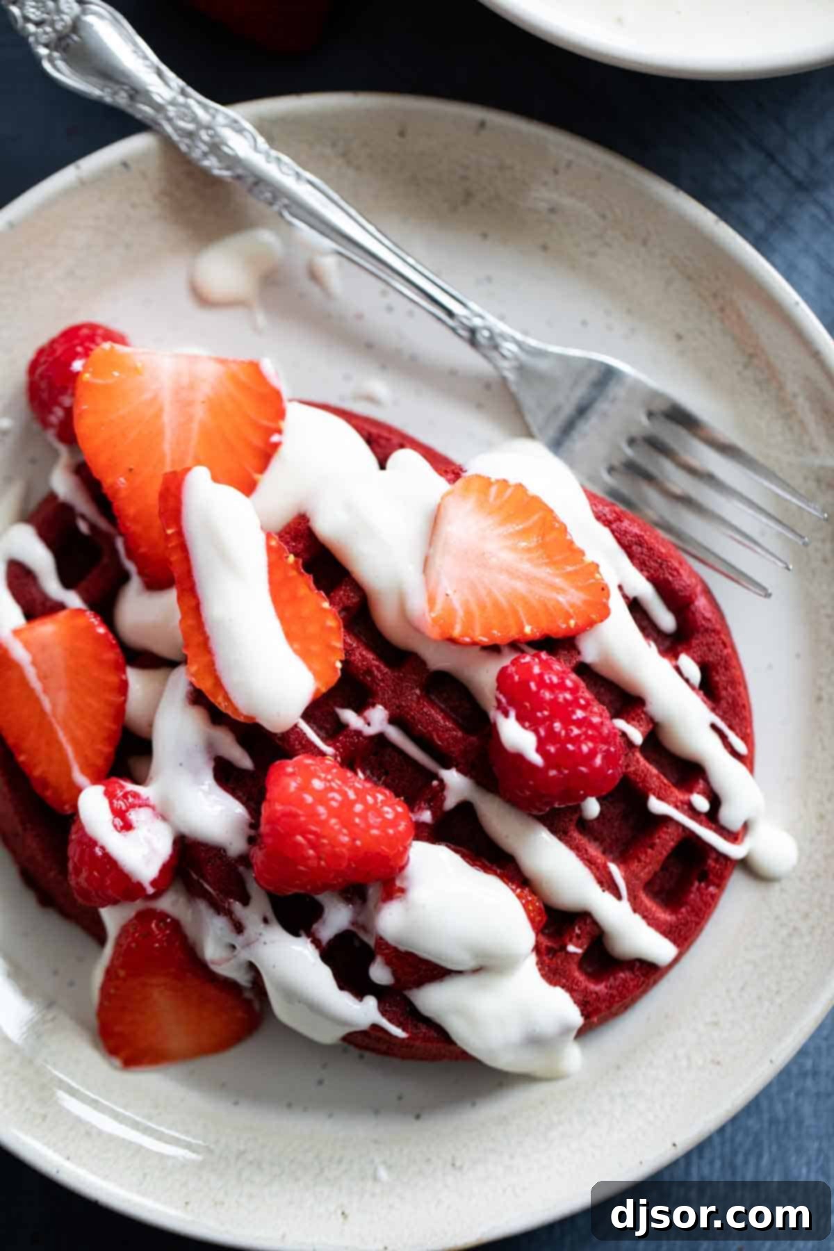 A stack of two beautifully cooked red velvet waffles, generously garnished with mixed berries and a delicate cream cheese drizzle.