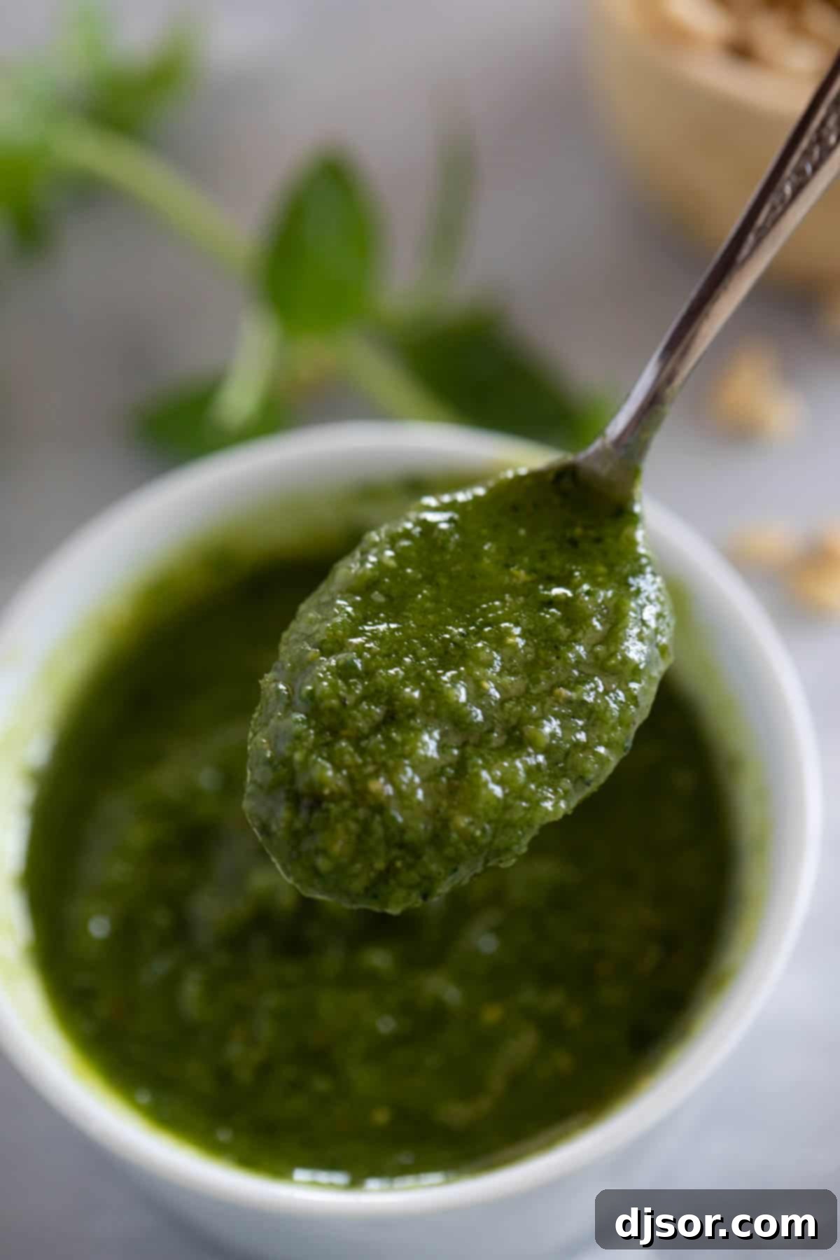 Fresh Basil Pesto 7 A spoon delicately holds a generous dollop of vibrant green basil pesto, positioned over a simple bowl, highlighting its rich and inviting texture.