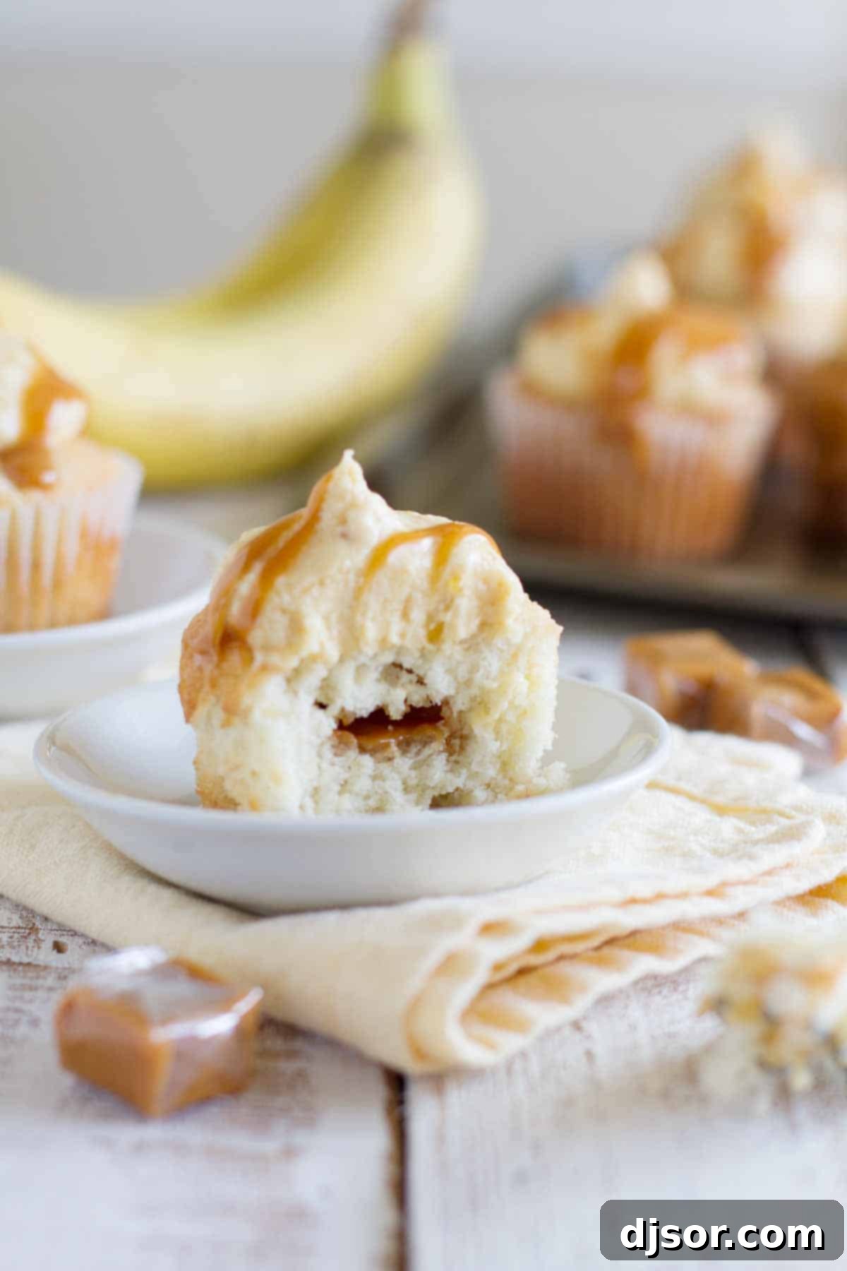 A close-up of a Caramel Banana Cupcake with a bite taken out, revealing the soft banana cake and the creamy, golden buttercream frosting.