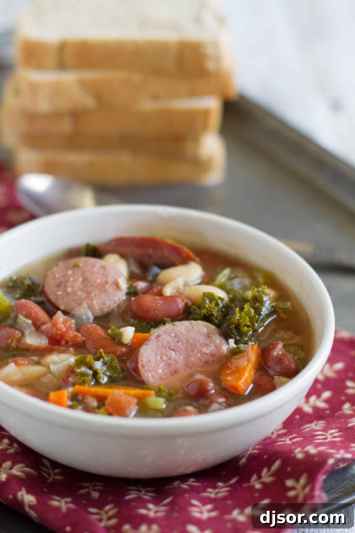 A steaming bowl of healthy Slow Cooker Tuscan Bean Soup with Sausage, featuring visible turkey sausage slices, kidney and great northern beans, and vibrant kale, carrots, and celery.