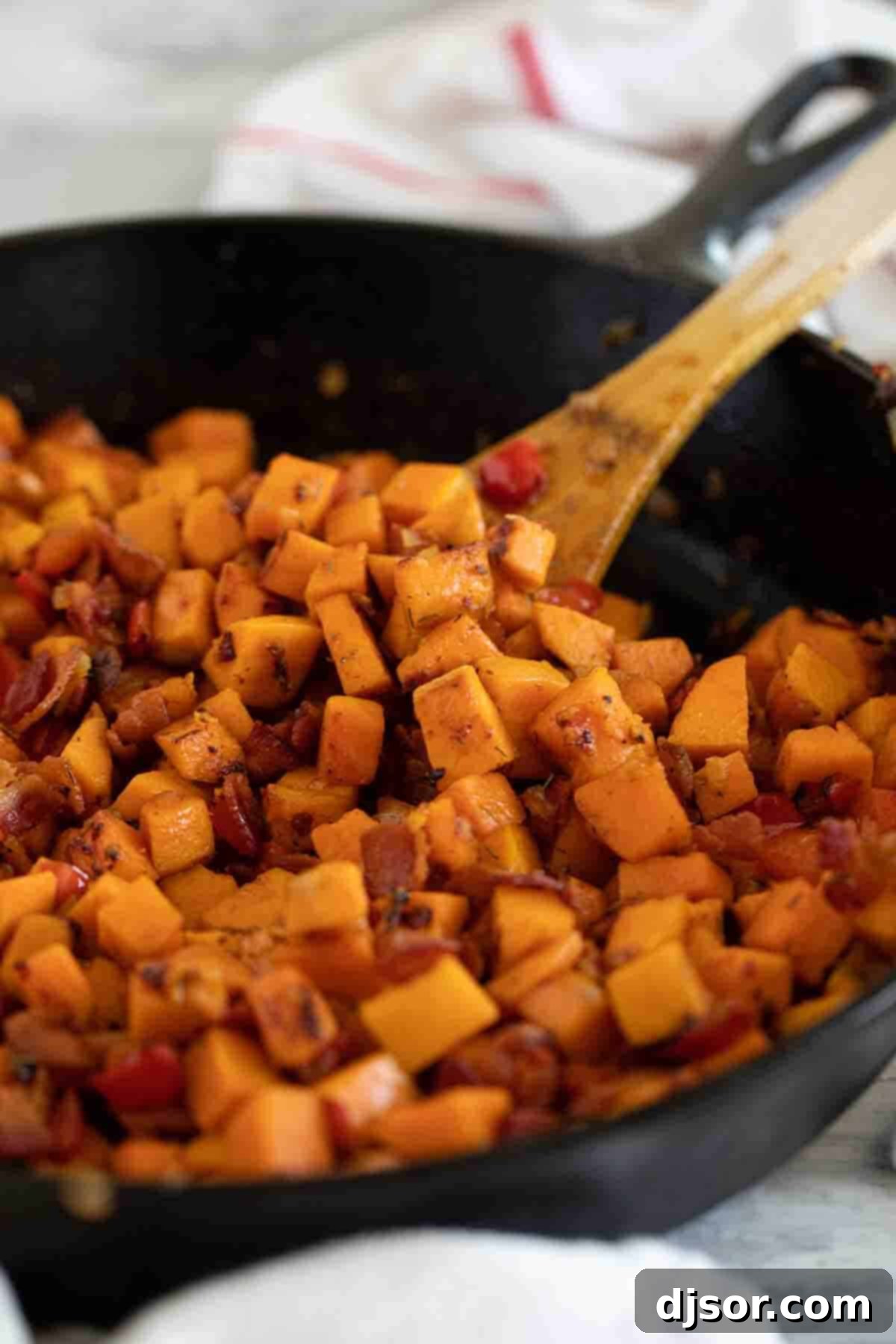 The perfect Sweet Potato Hash – sweet potatoes are combined with bacon, onions and sweet red peppers in this easy stovetop dish. Serve it as a side dish or add an egg on top and turn it into breakfast. A wooden spoon scooping into a freshly made skillet of Sweet Potato Hash, highlighting its rich textures and vibrant colors.
