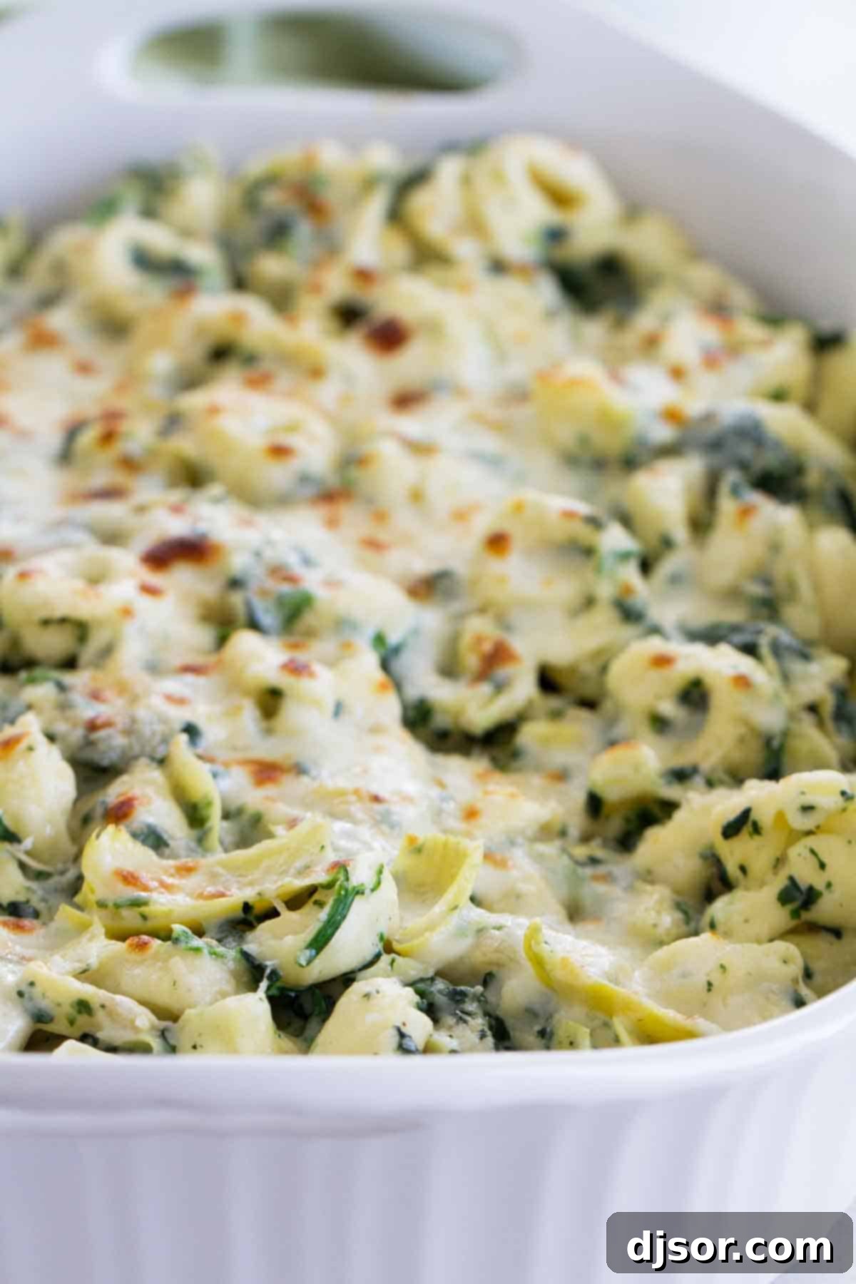 Close-up of a white baking dish showcasing the golden, bubbly cheese on top of the Spinach and Artichoke Tortellini Bake, fresh from the oven.