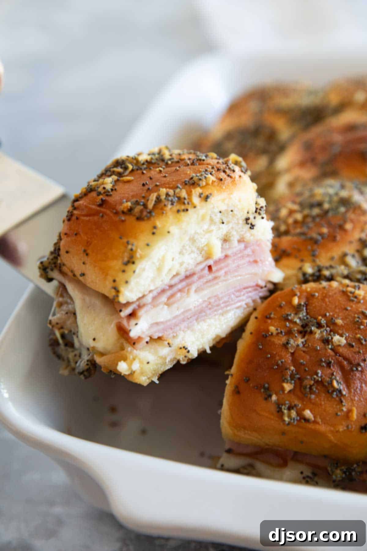 A hand reaching into a casserole dish to pick up a single golden-brown Ham and Cheese Slider, showcasing its readiness to be enjoyed.