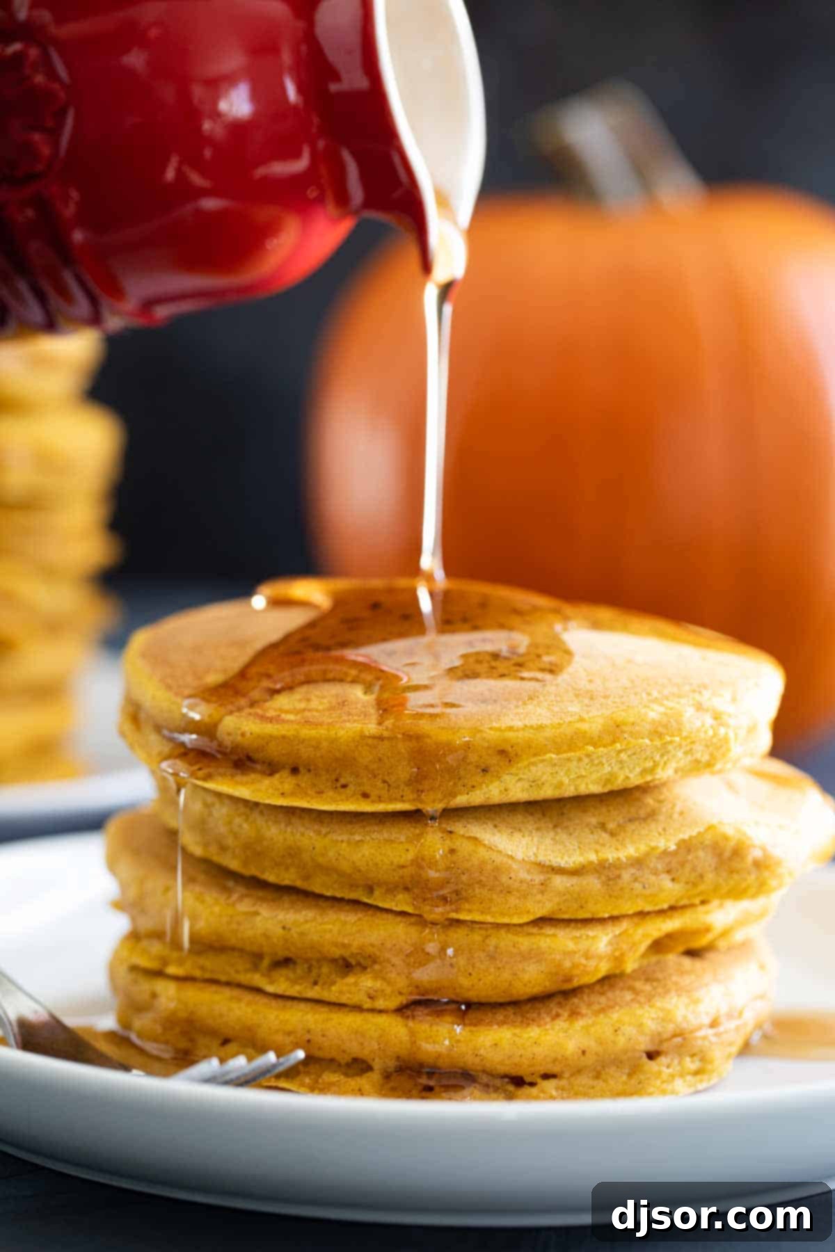 Maple syrup being poured over a stack of warm pumpkin pancakes, highlighting their fluffy texture and golden-brown edges.