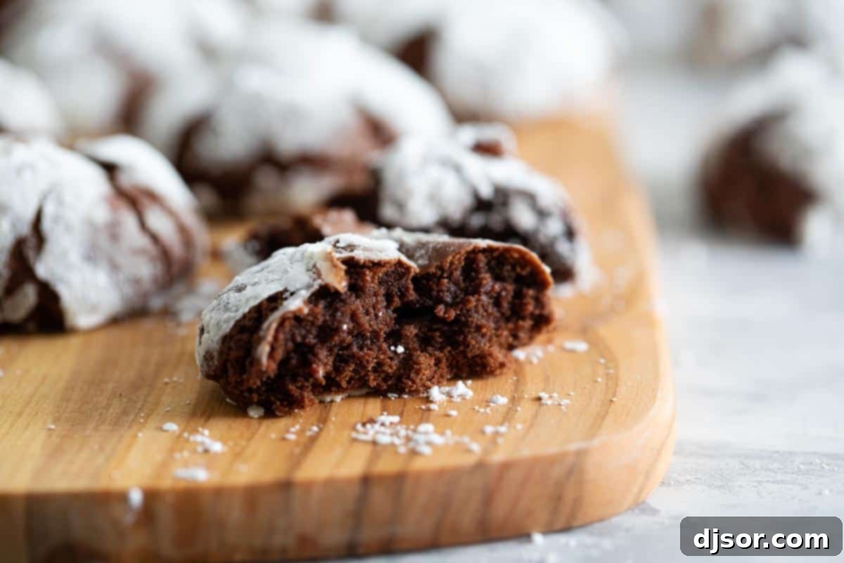 A chocolate crinkle cookie broken in half, revealing its moist, fudgy, brownie-like interior and contrasting with the crisp, powdered sugar exterior.