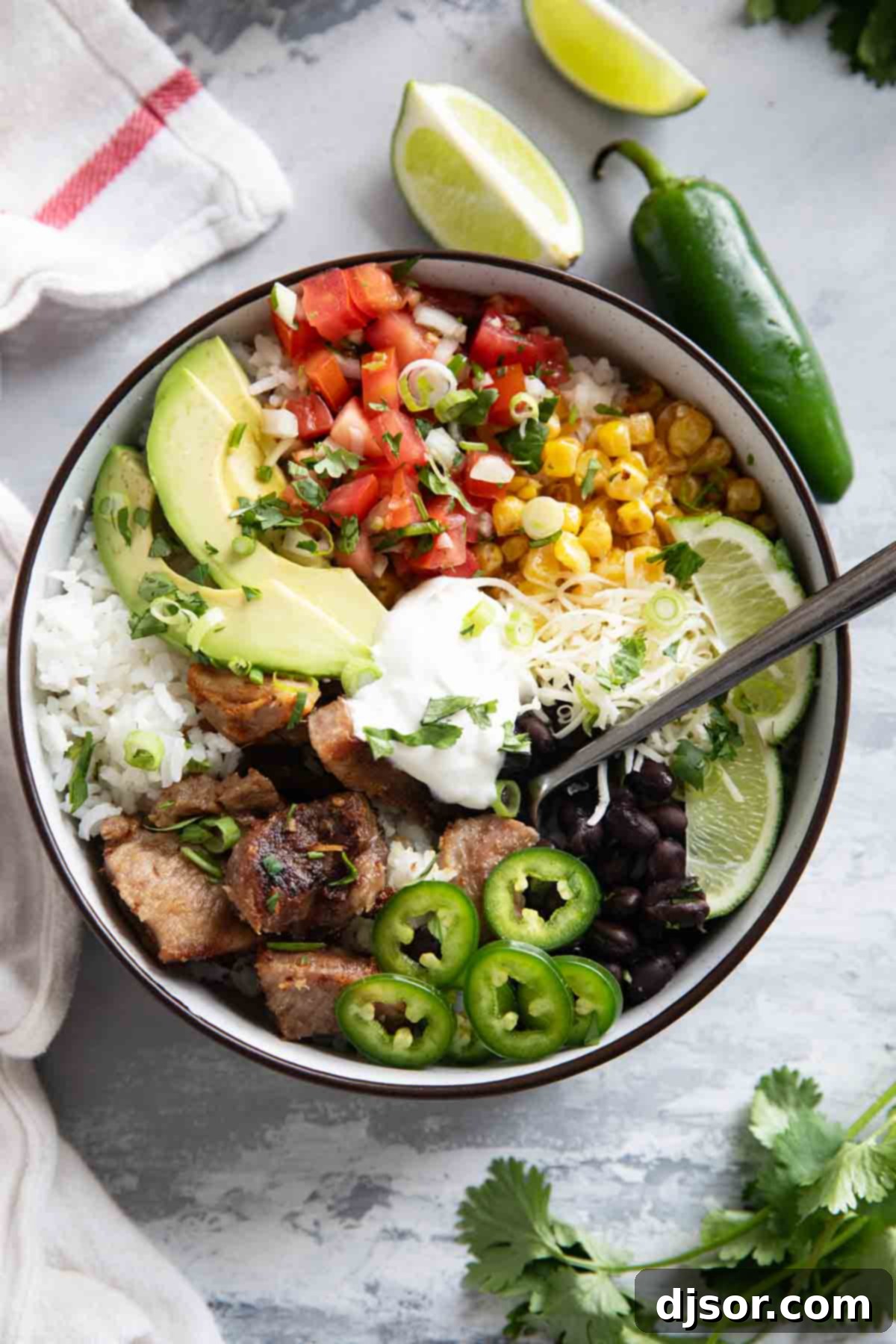 A vibrant Pork Carnitas Burrito Bowl, generously topped with crisp jalapeños, fresh pico de gallo, shredded cheese, creamy avocado slices, hearty black beans, a dollop of sour cream, and tender, caramelized pork carnitas.